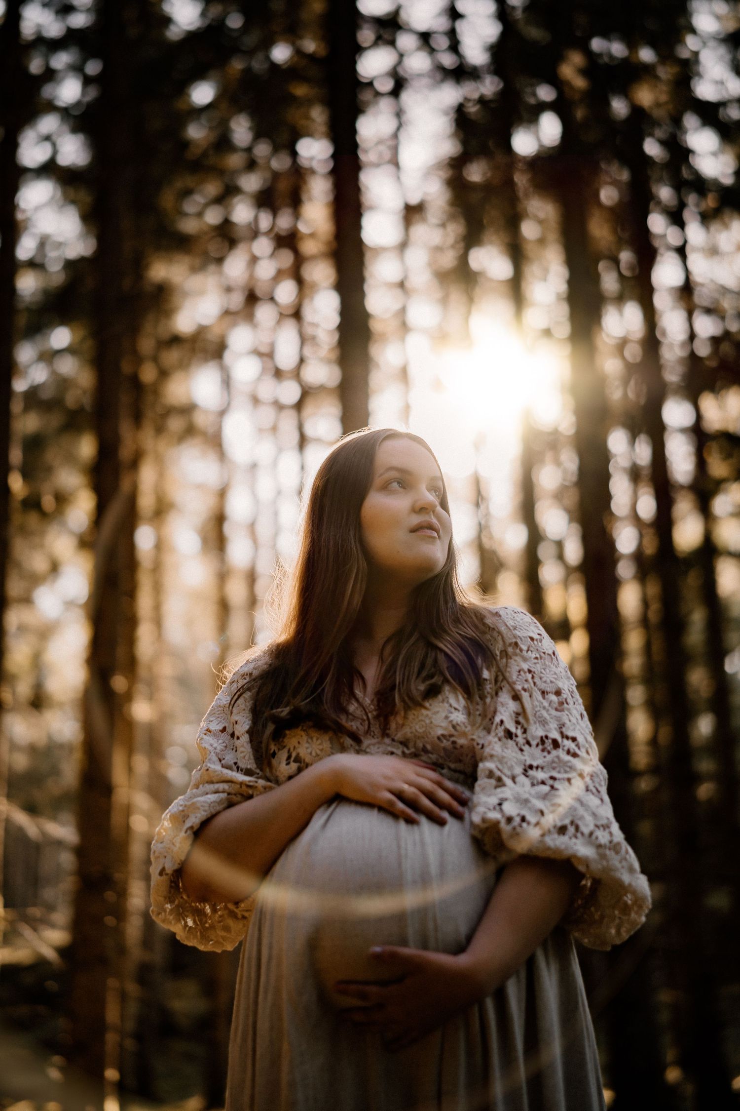 A dreamy maternity photo in a sunlit forest with soft bokeh and a woman wearing a lace shawl over a flowing dress.