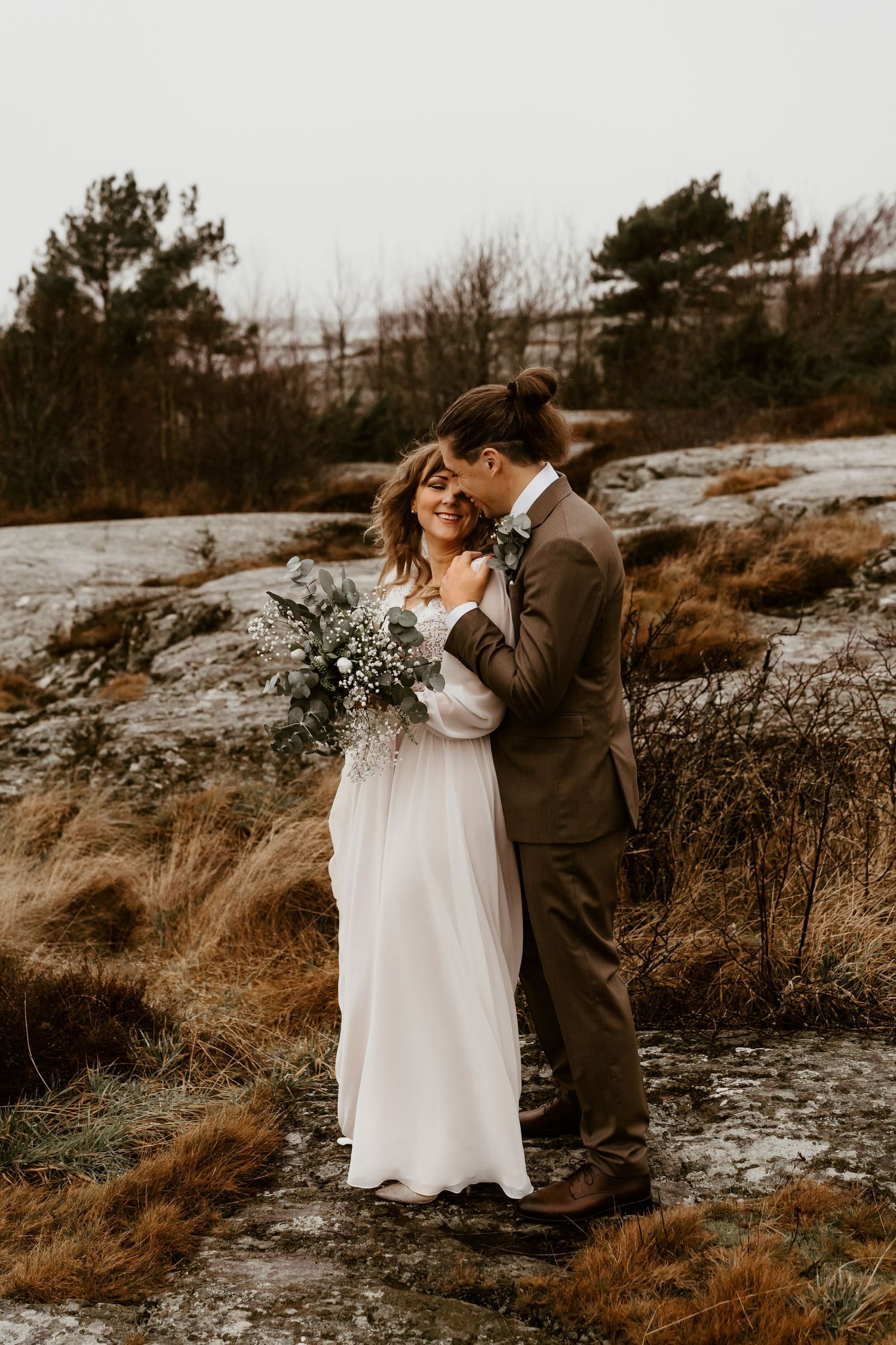 A couple in wedding attire embrace on rocky terrain during a moody autumn sunset photoshoot.