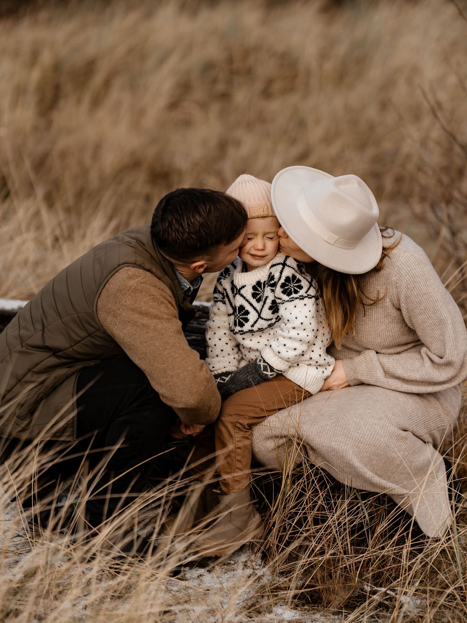 Family sharing tender moment together on a field of dry grass wearing neutral toned clothing.
