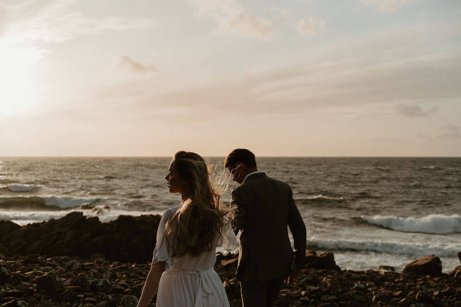 Silhouettes of a couple in wedding attire stand on rocky coastline at sunset as ocean waves crash behind them.
