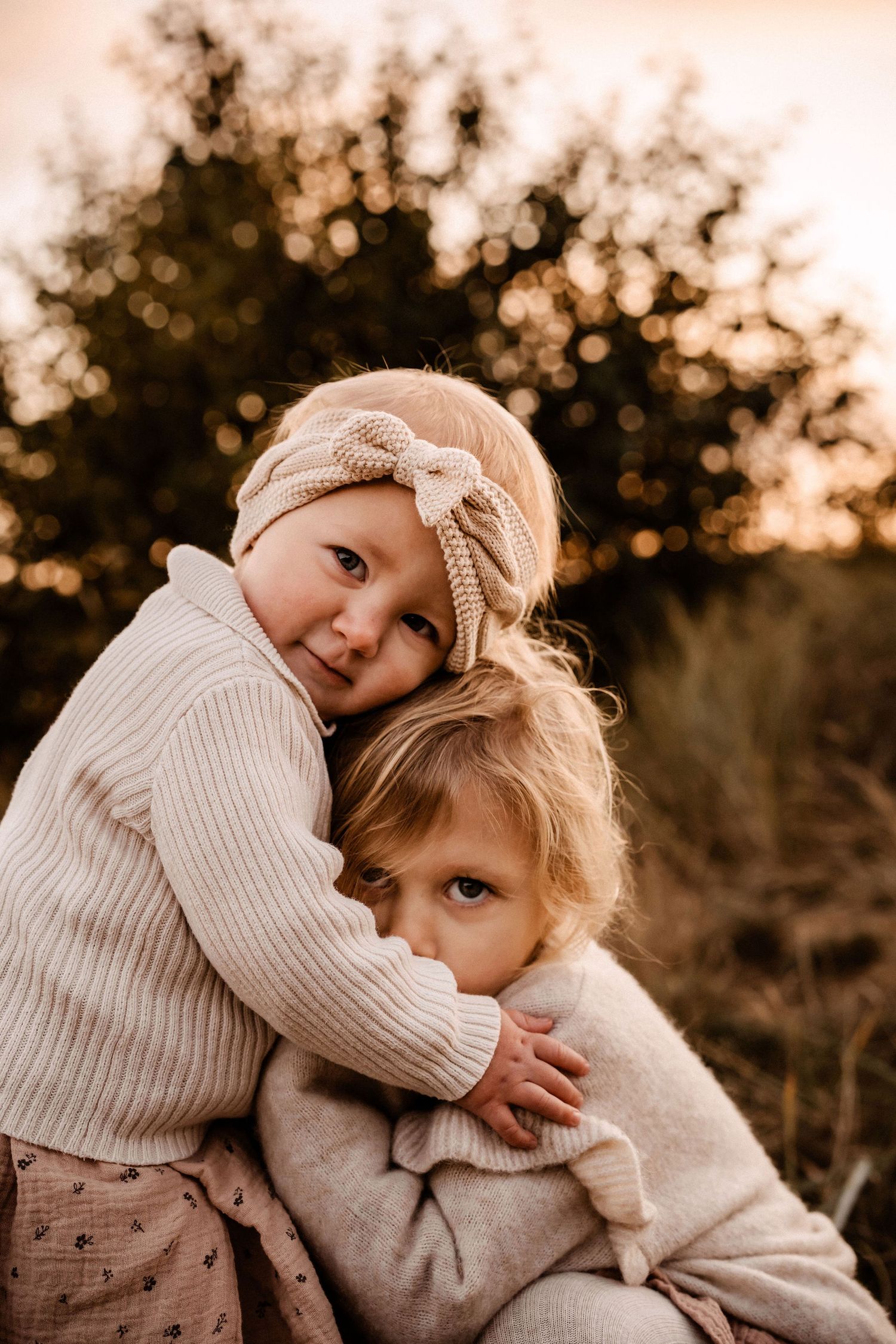 Two children share a sweet embrace while wearing cozy knit sweaters in warm sunset lighting.