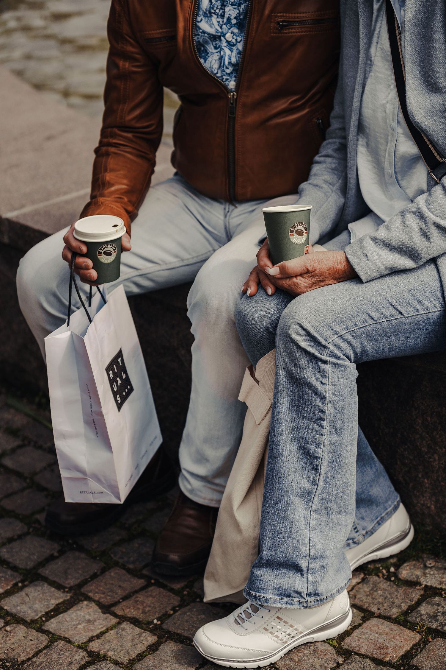 Two people in casual attire sitting with coffee cups and shopping bags on a cobblestone street.