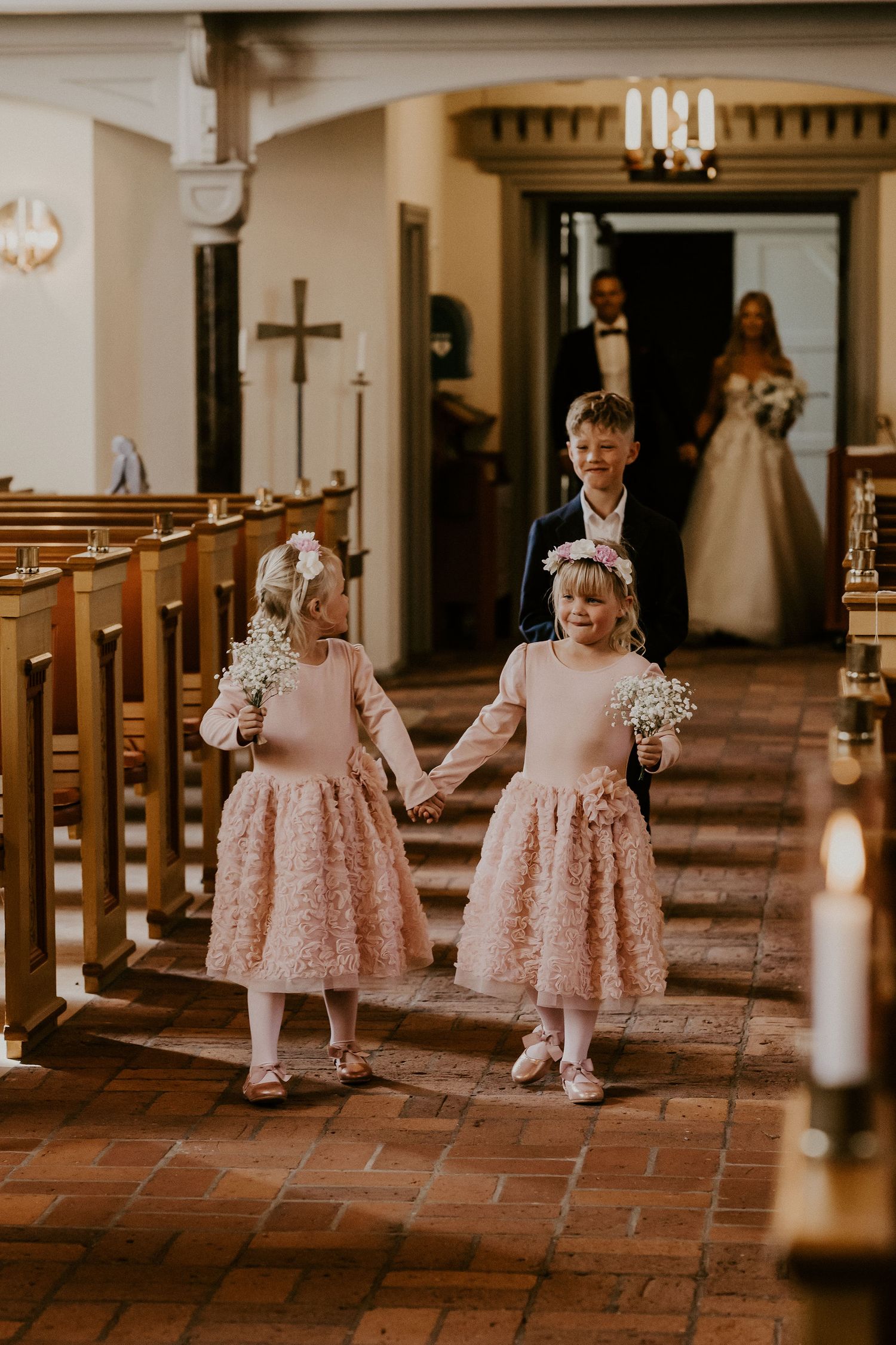 Two young flower girls in matching pink lace dresses walk down the church aisle holding hands.