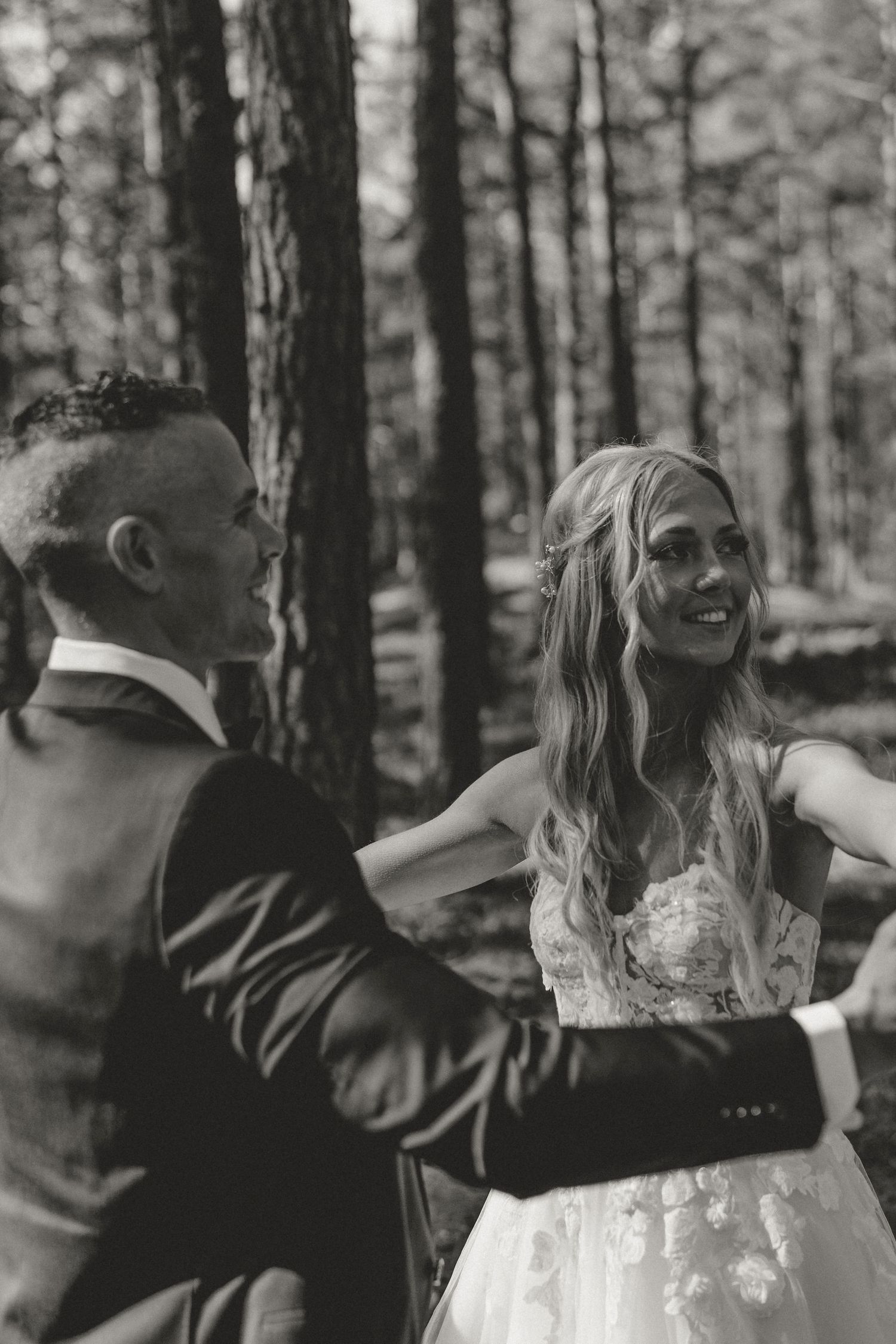 A couple in a forest setting sharing a joyful moment as the bride in a white lace dress laughs candidly.