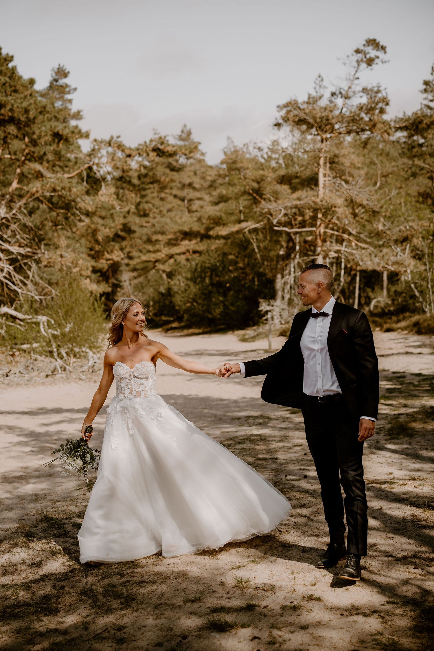 A couple in formal wedding attire holds hands and smiles while standing on a path surrounded by pine trees.