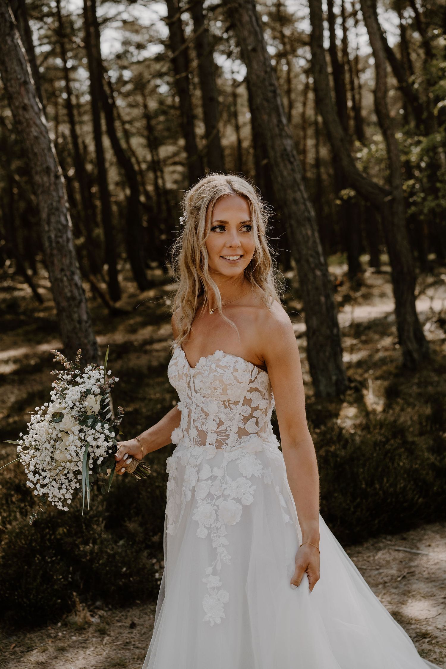 A bride in a white lace wedding dress poses in a wooded forest setting holding a bouquet of baby's breath.
