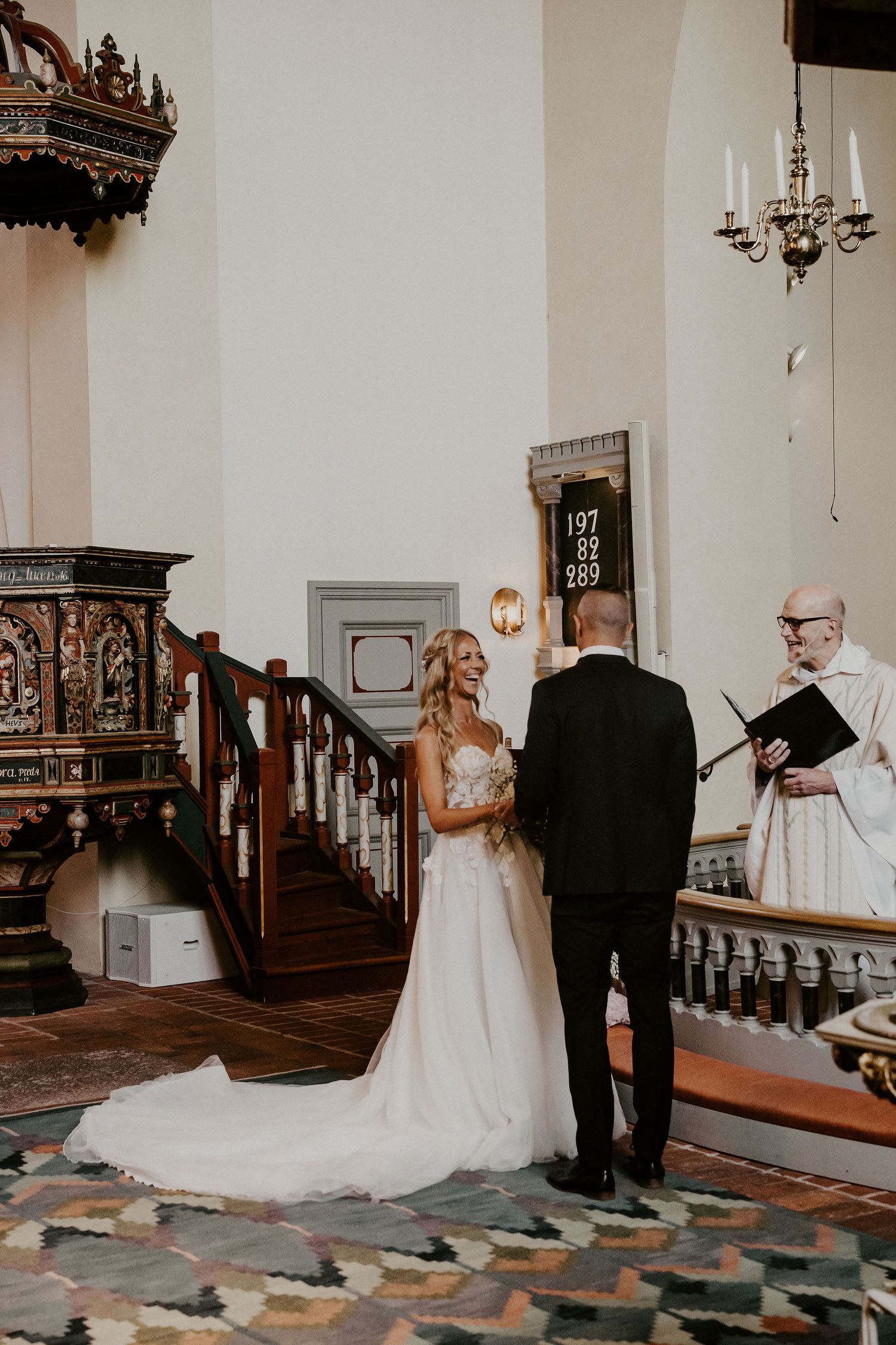 A wedding ceremony takes place in a traditional church with ornate wooden details and a chandelier.