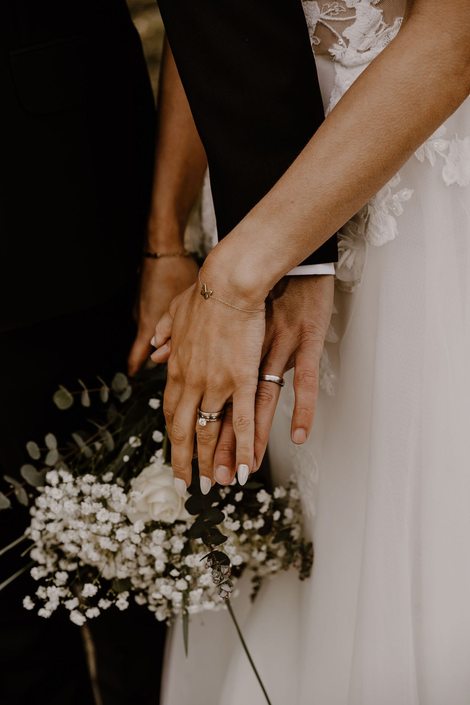 Close-up of newlyweds' hands with wedding rings holding white baby's breath flowers against black and white attire.