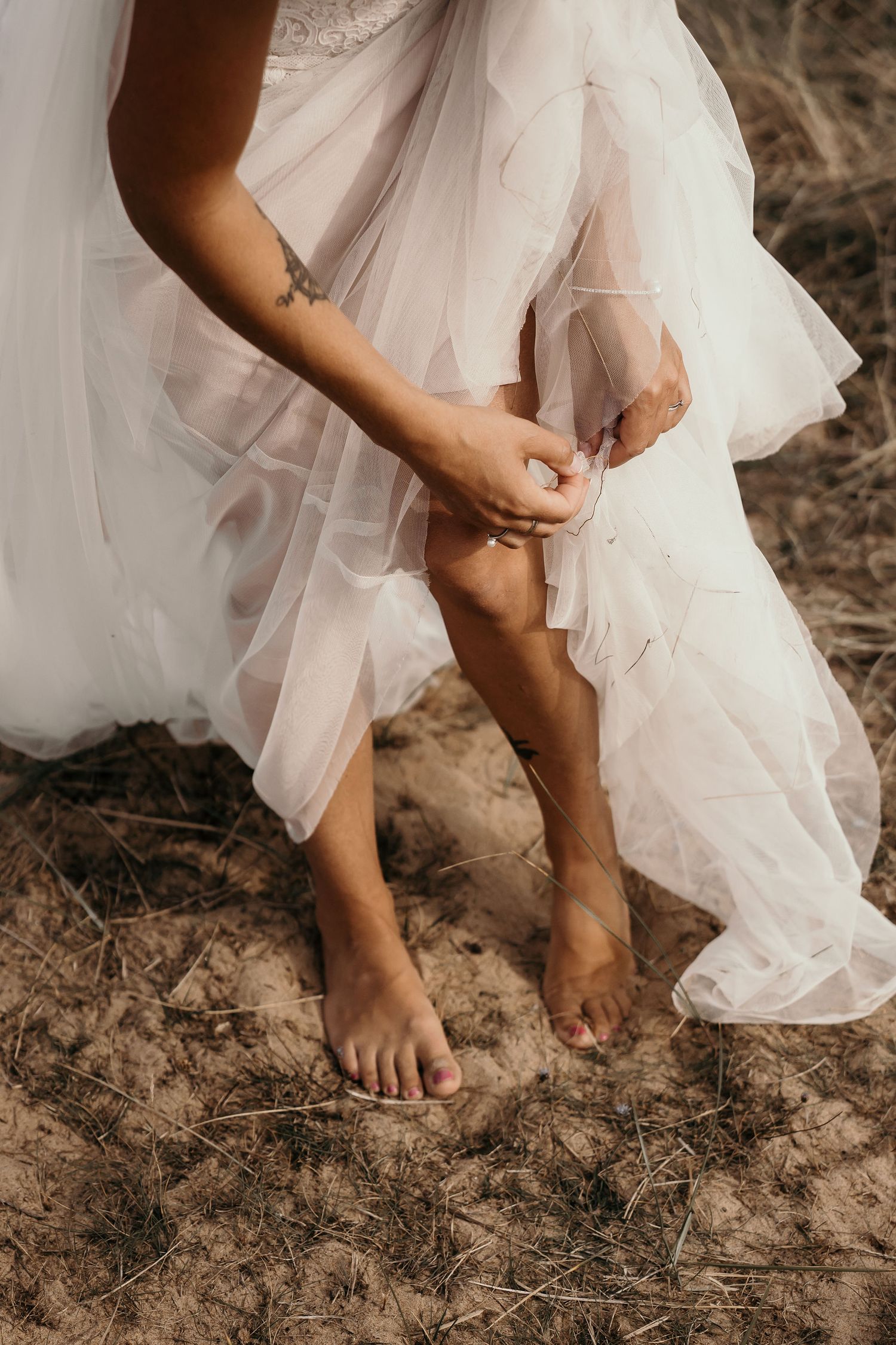 Bride in flowing tulle dress adjusts her anklet while standing barefoot on rustic ground.