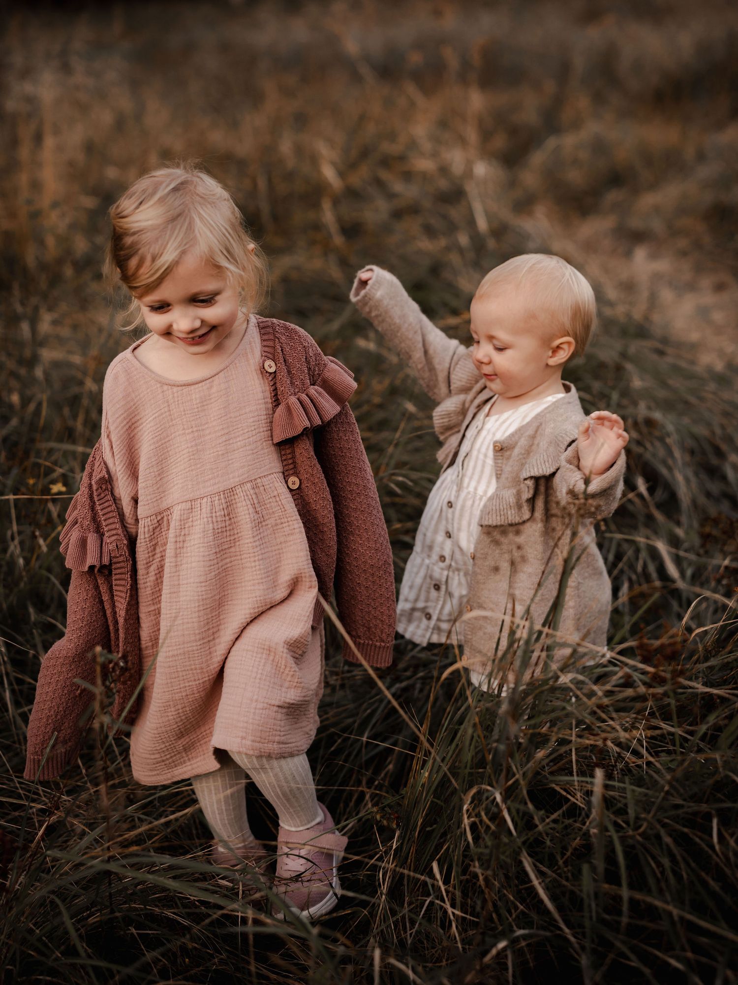 Two young children in muted earth-toned dresses walking together through tall brown grass at sunset.