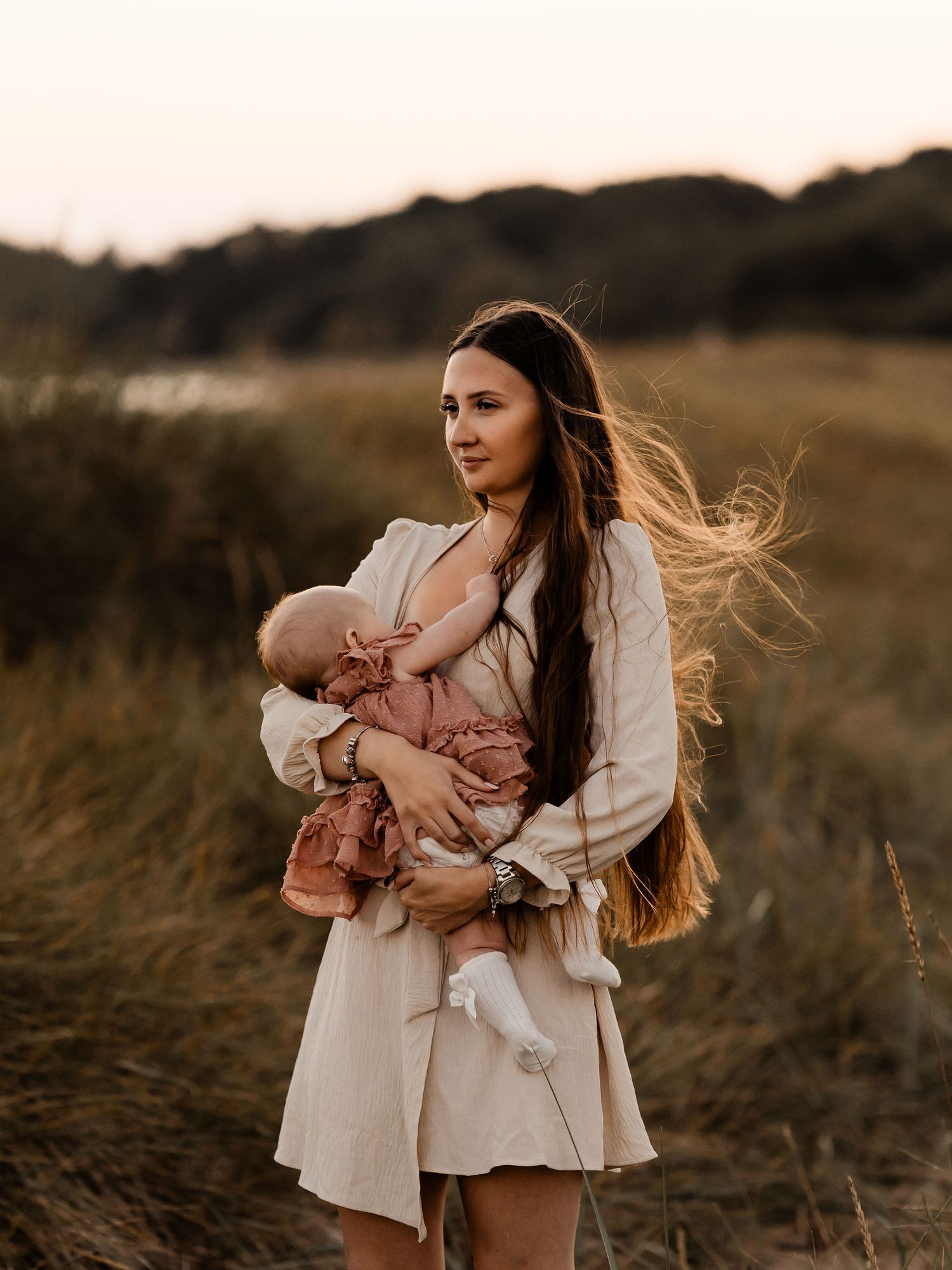 Long-haired woman in cream dress holds sleeping infant against warm sunset backdrop in grassy field.