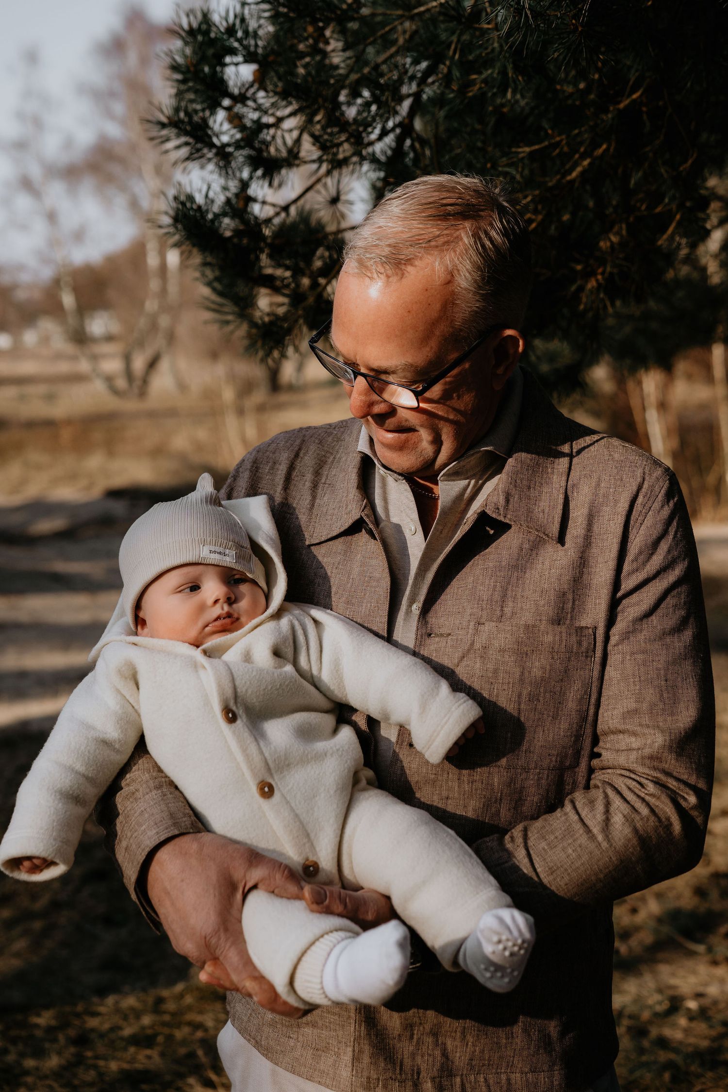 A person wearing glasses and brown jacket holds a small baby bundled in white clothing outdoors on a sunny day.