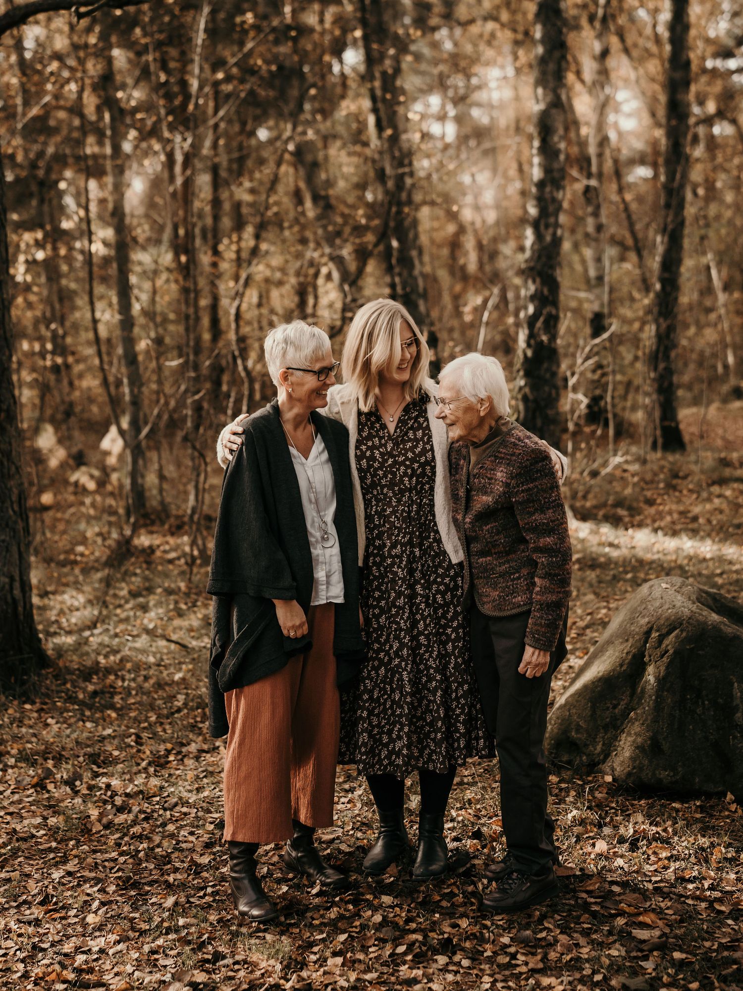 Three generations stand together on a leaf-covered forest path during autumn, wearing warm layers in muted earth tones.