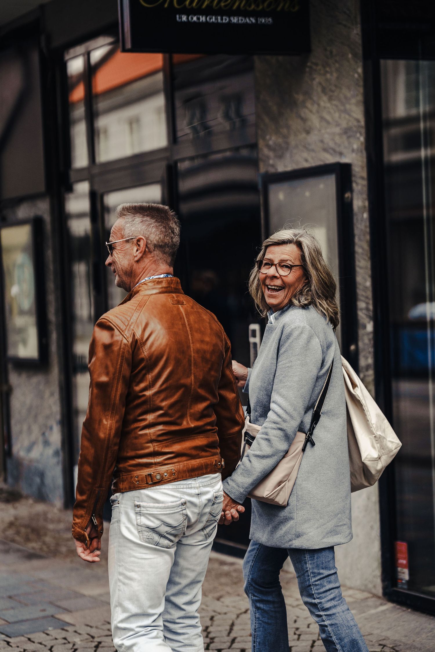 Stylish couple walking down urban street, laughing and smiling while wearing casual leather jacket and gray coat.