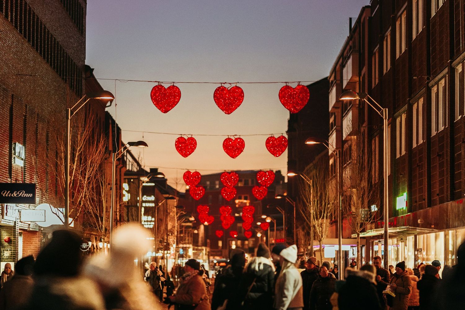 Red heart-shaped lanterns illuminate a bustling city street at dusk during a festive celebration.