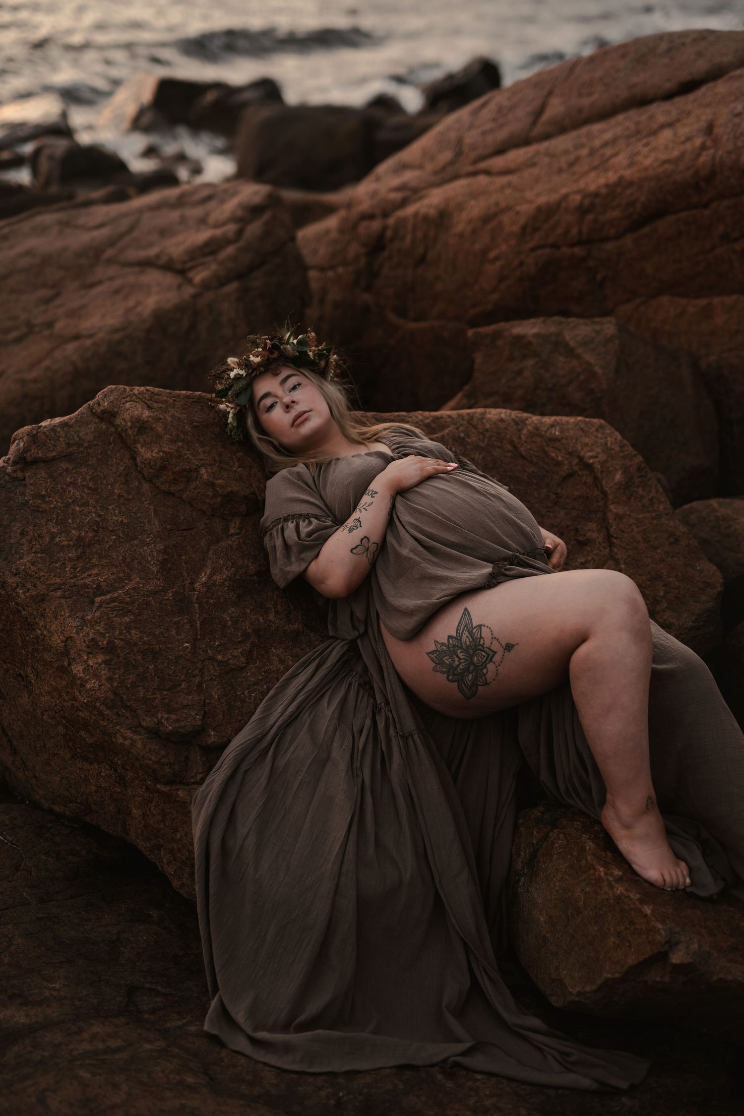 Artistic maternity photo on rocky beach featuring flowing brown dress and floral crown at sunset.