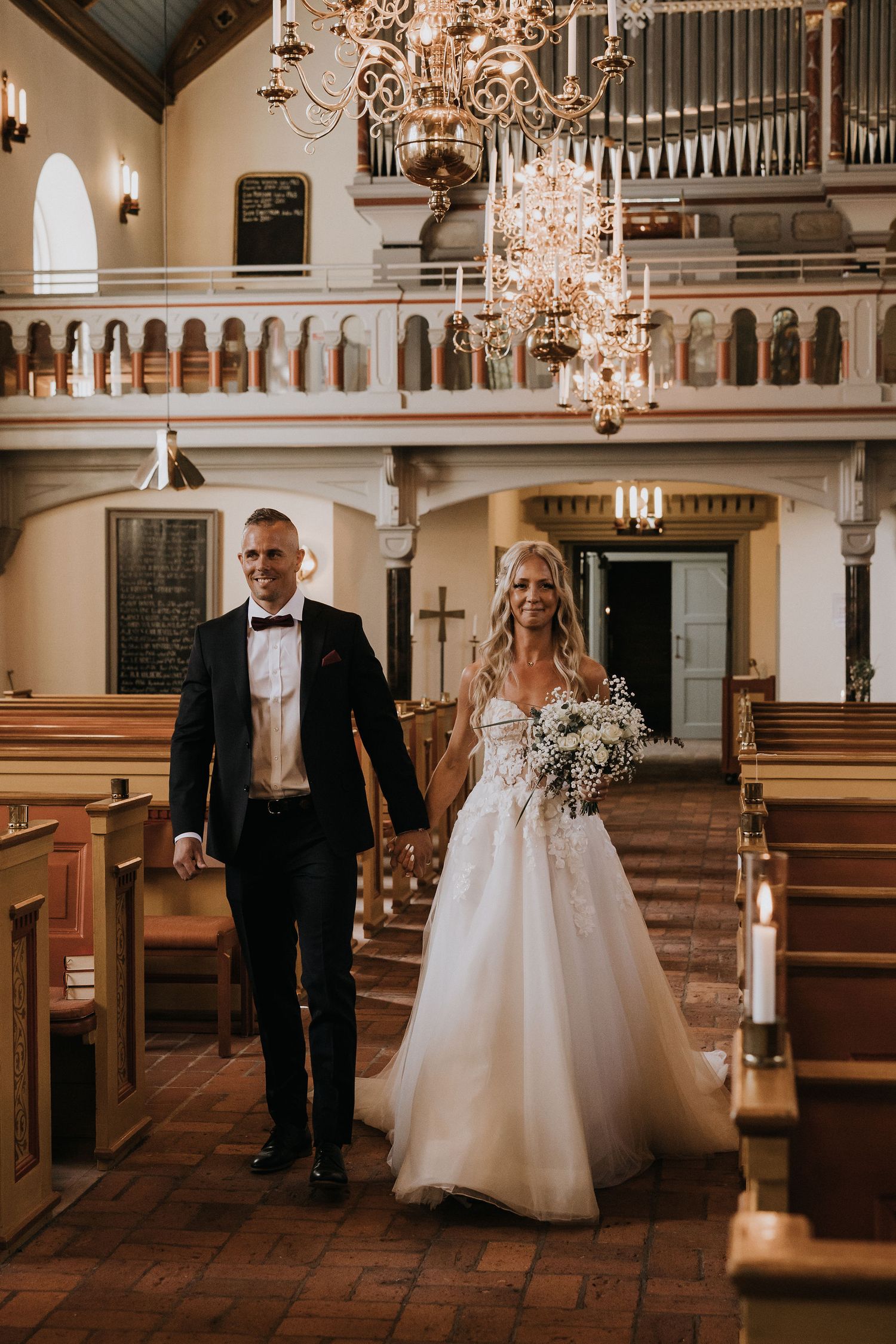A couple walks down the aisle of a historic church with elegant chandeliers and organ pipes in the balcony.