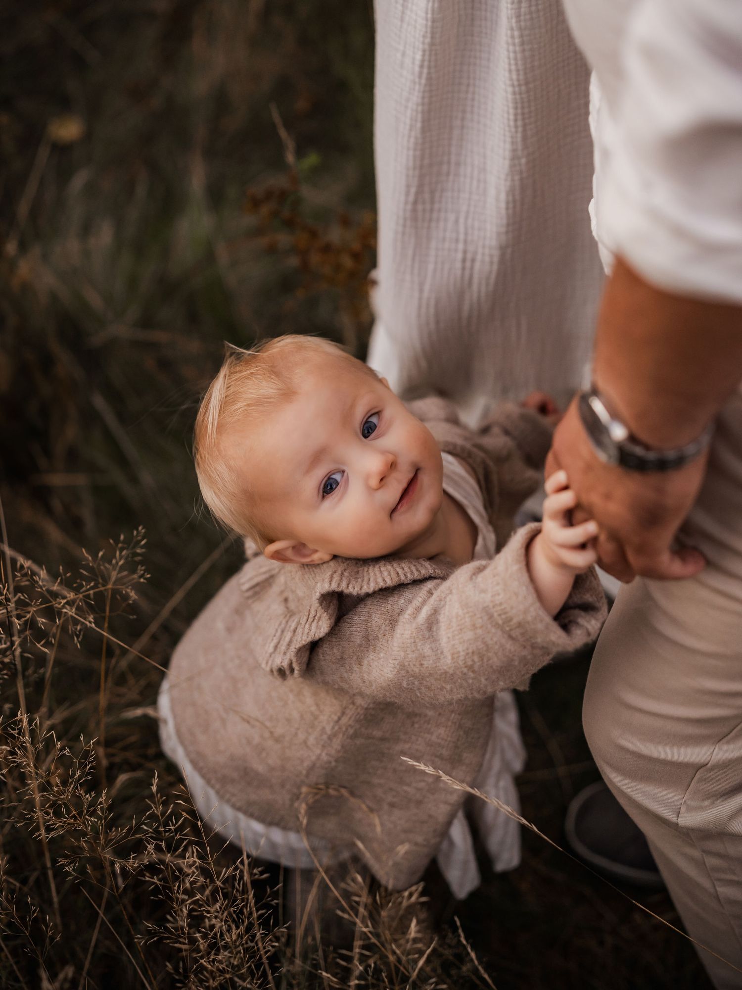 Small child in knit sweater smiles while holding onto parent's leg in outdoor setting.