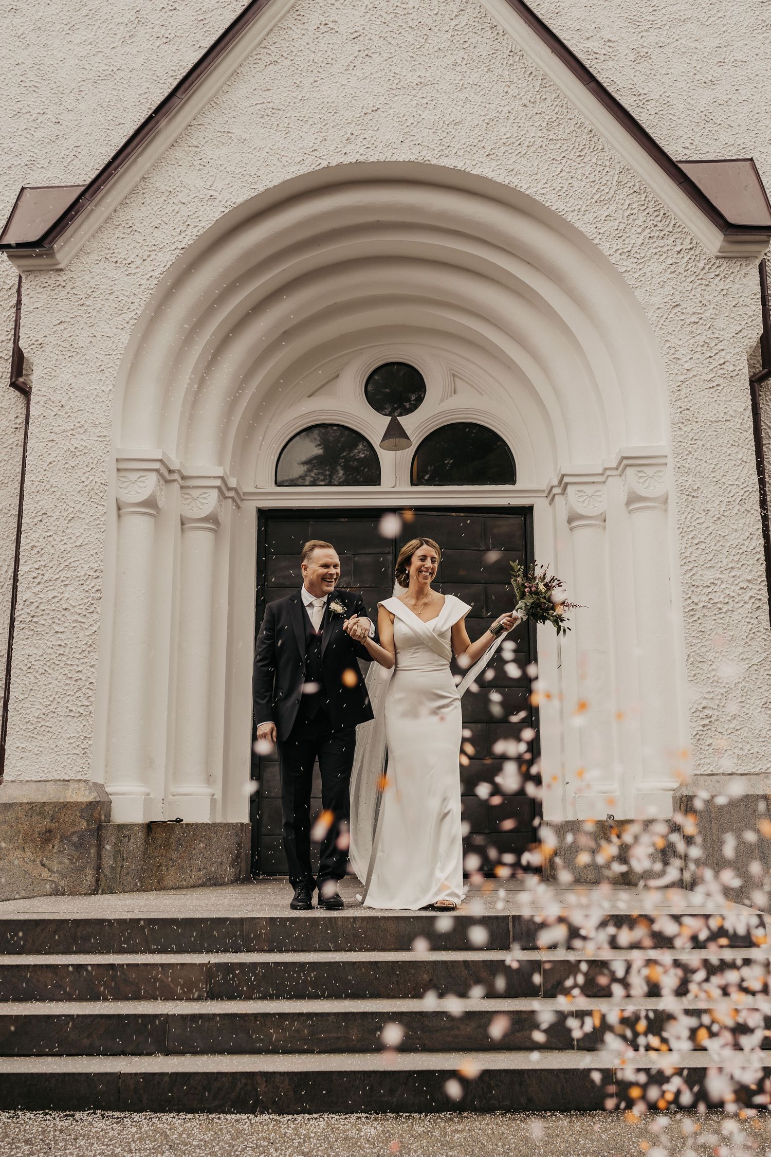 Two people exit a white church with an arched doorway as confetti falls during a wedding celebration.