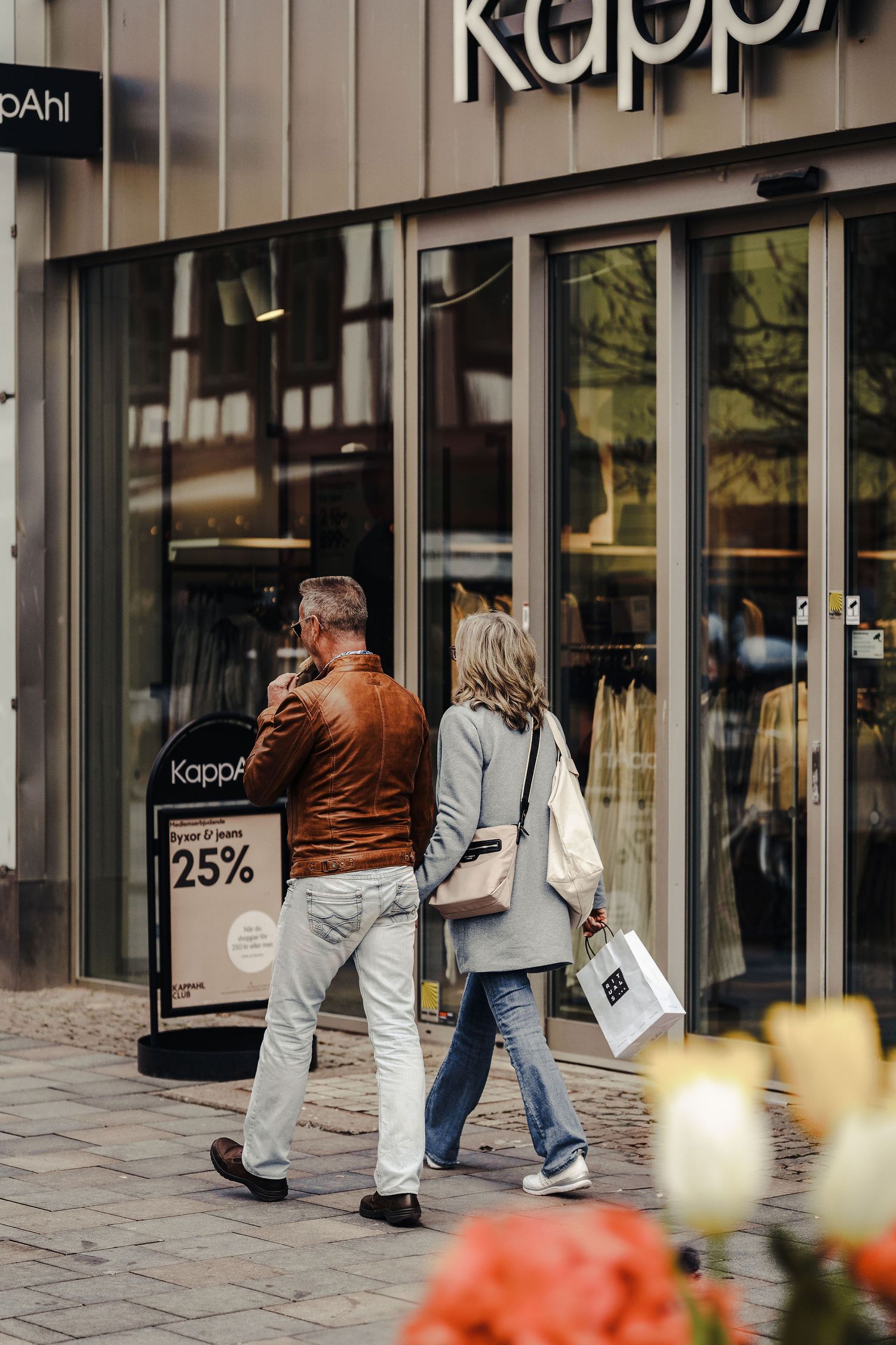 Two shoppers walk towards a Kappahl store entrance advertising 25 percent off sale on a city street.