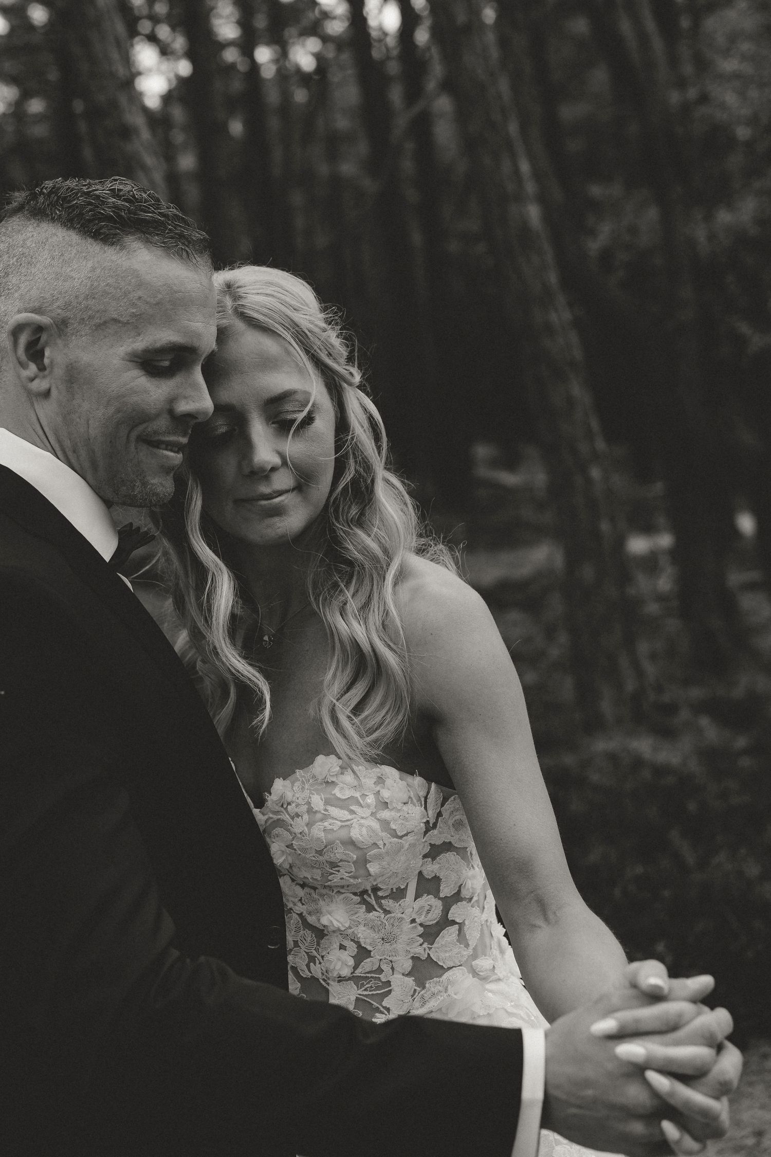 A romantic black and white wedding photo shows a couple in formal attire sharing an intimate moment in a wooded setting.