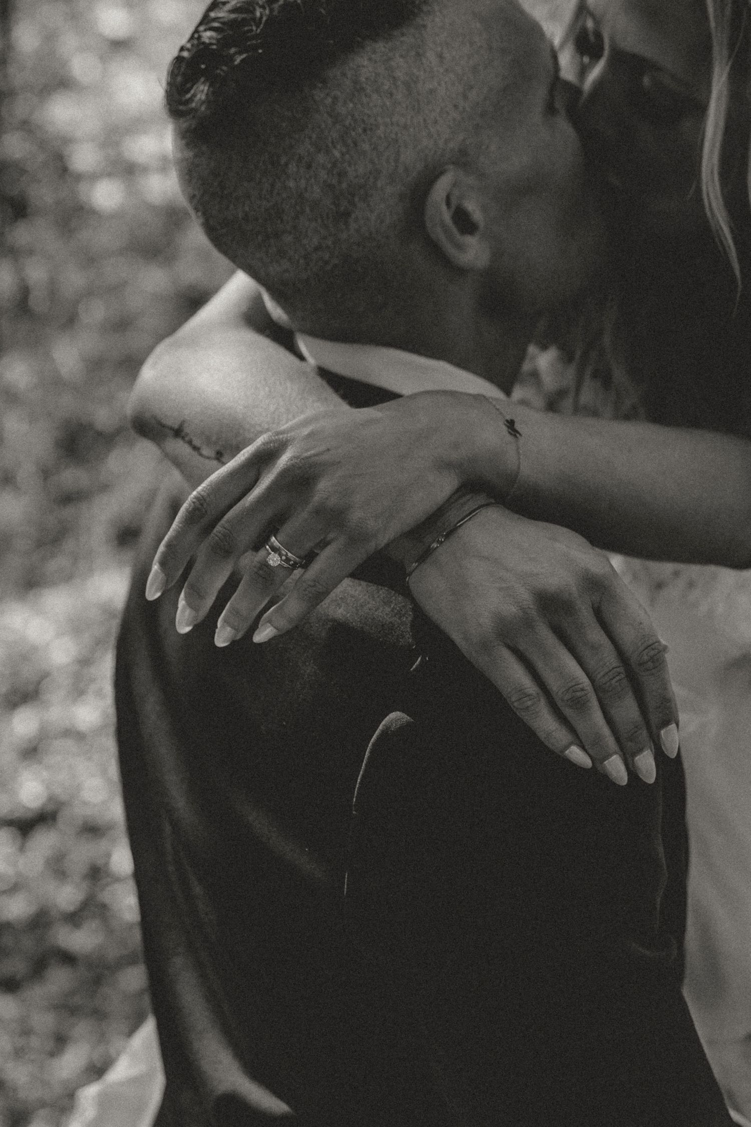 Black and white intimate photo showing arms embracing with an engagement ring visible on one hand.