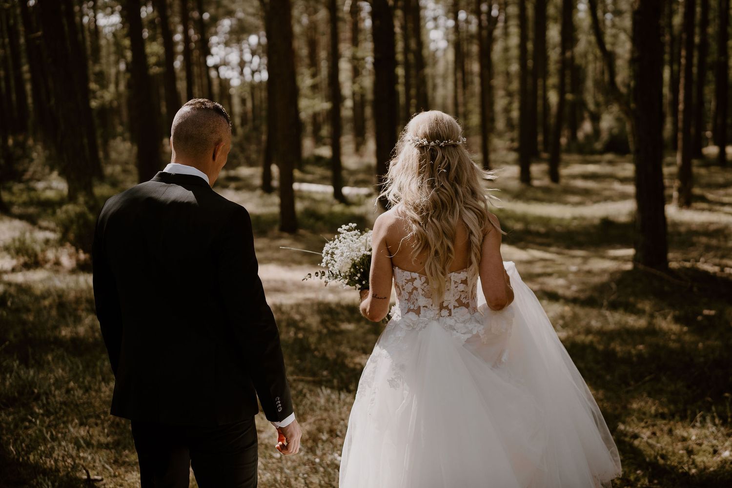 A couple walks hand in hand through a sunlit forest on their wedding day, with the bride in a flowing white gown.
