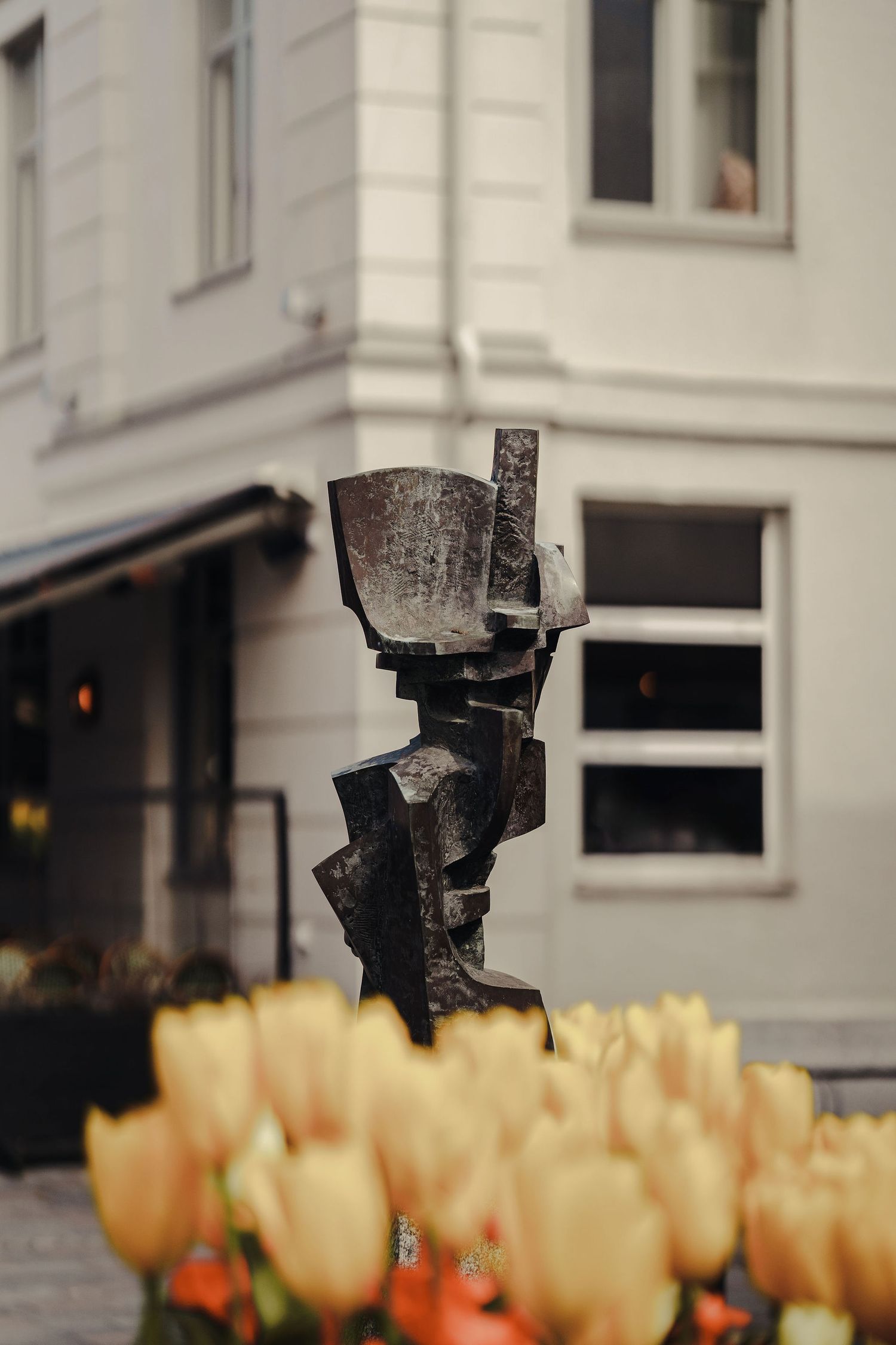 A rustic metal sculpture stands in front of a white building with orange tulips blooming in the foreground.
