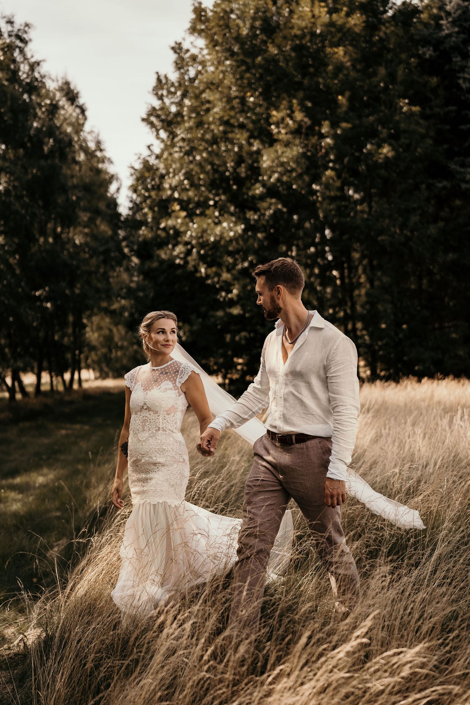 A couple in wedding attire walks together through tall grass at sunset in a romantic outdoor setting.