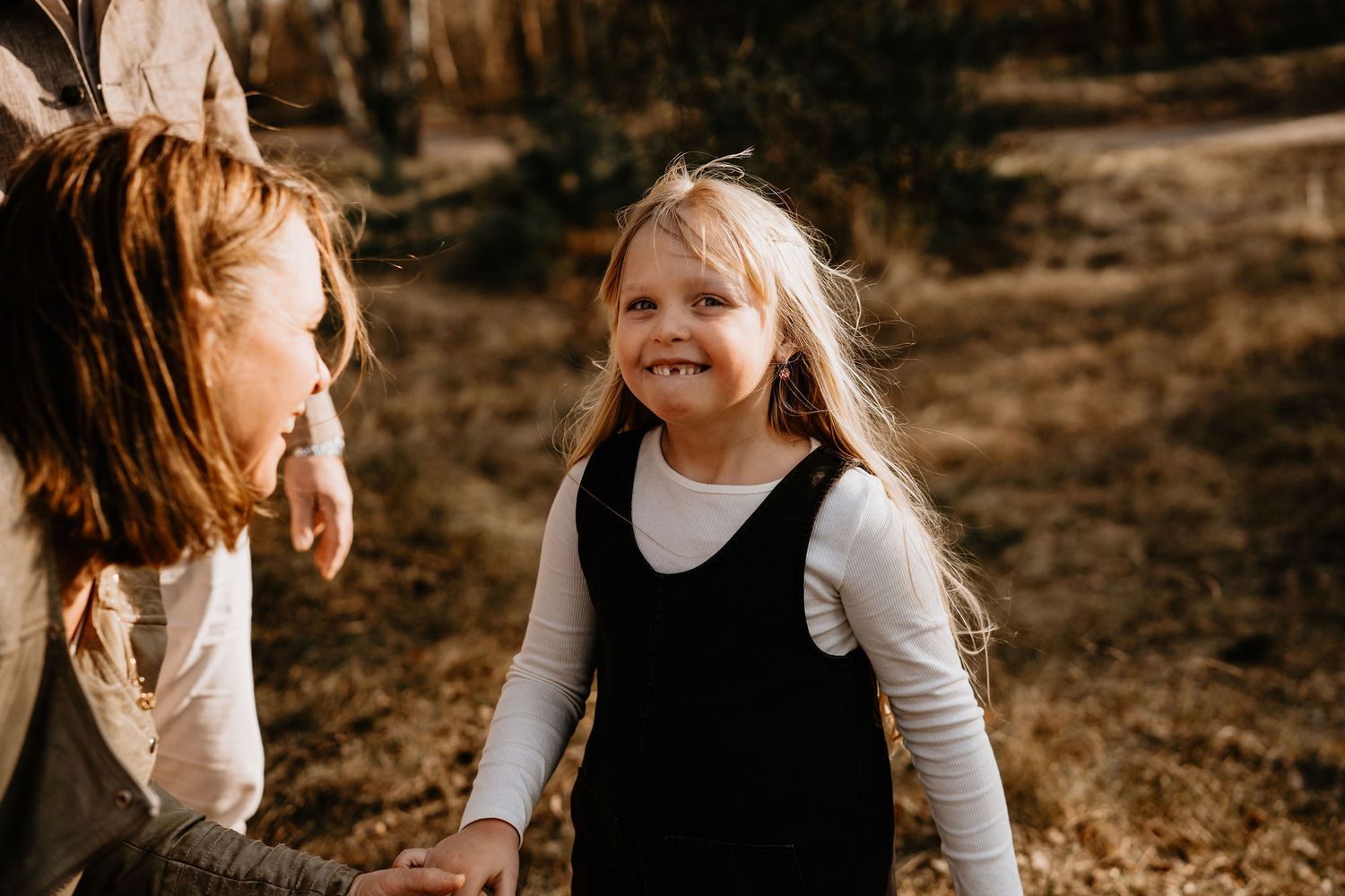 A happy child in a white shirt and black jumper dress stands outside in autumn sunlight while interacting with someone nearby.