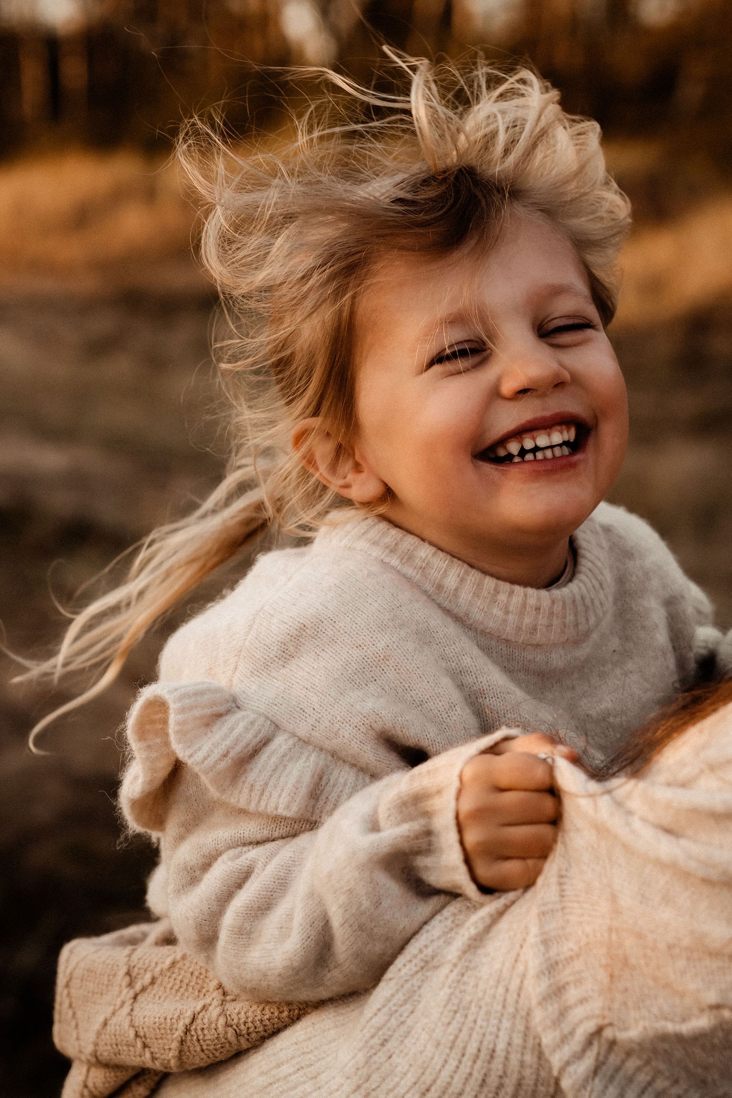 A laughing child with windswept hair wears a cream-colored knit sweater in golden sunlight.