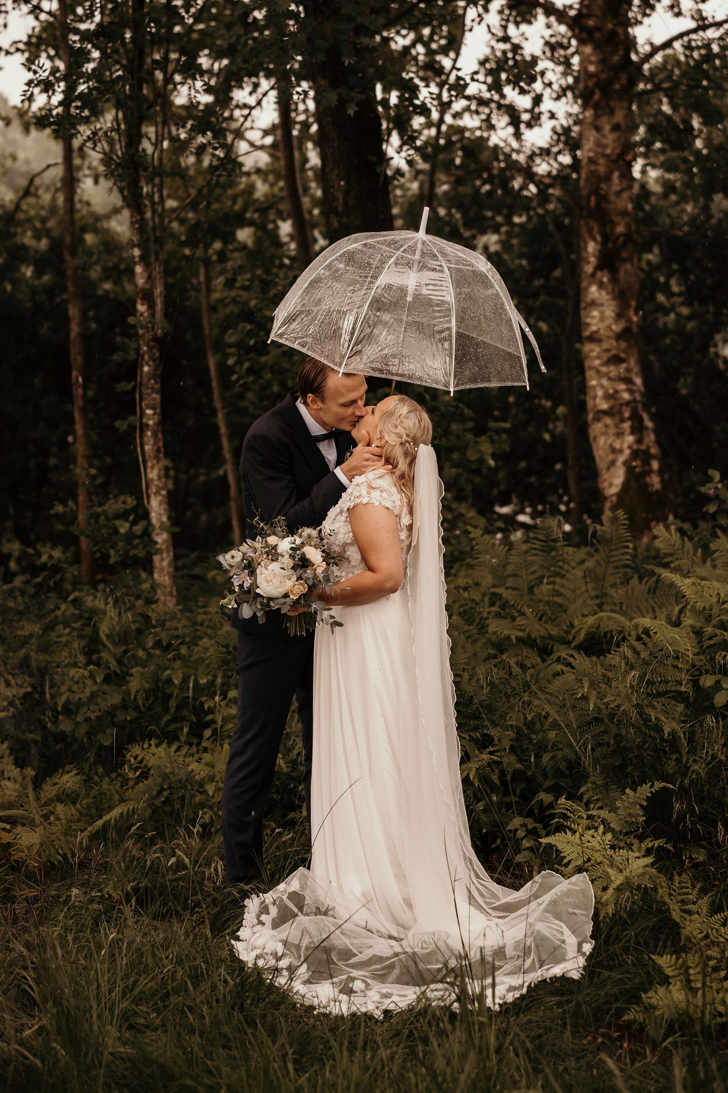 A romantic wedding couple shares a moment under a clear umbrella in a moody forest setting with elegant flowers.