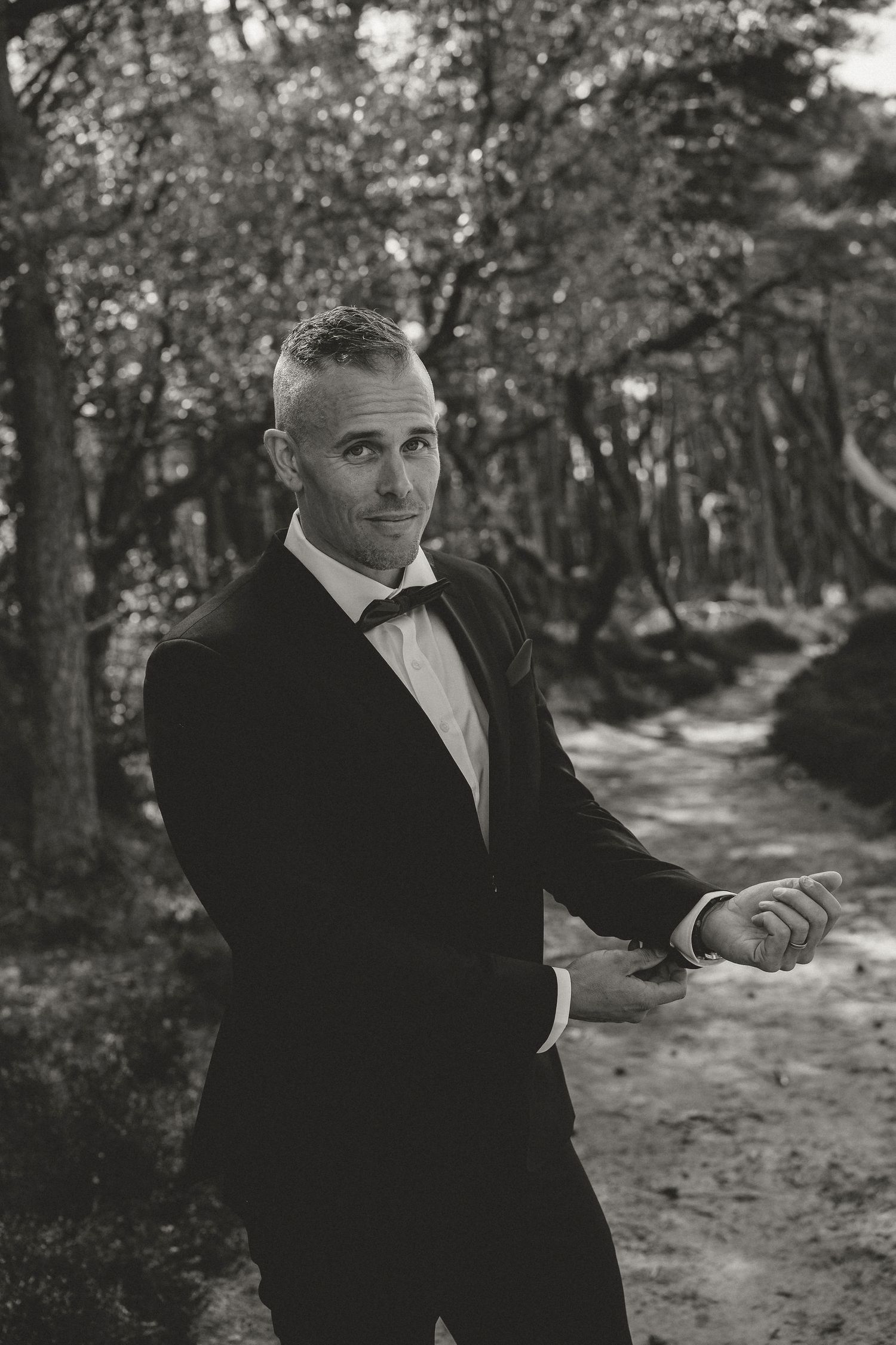 Black and white photo of a groom in a classic tuxedo and bowtie posing on a wooded garden path.