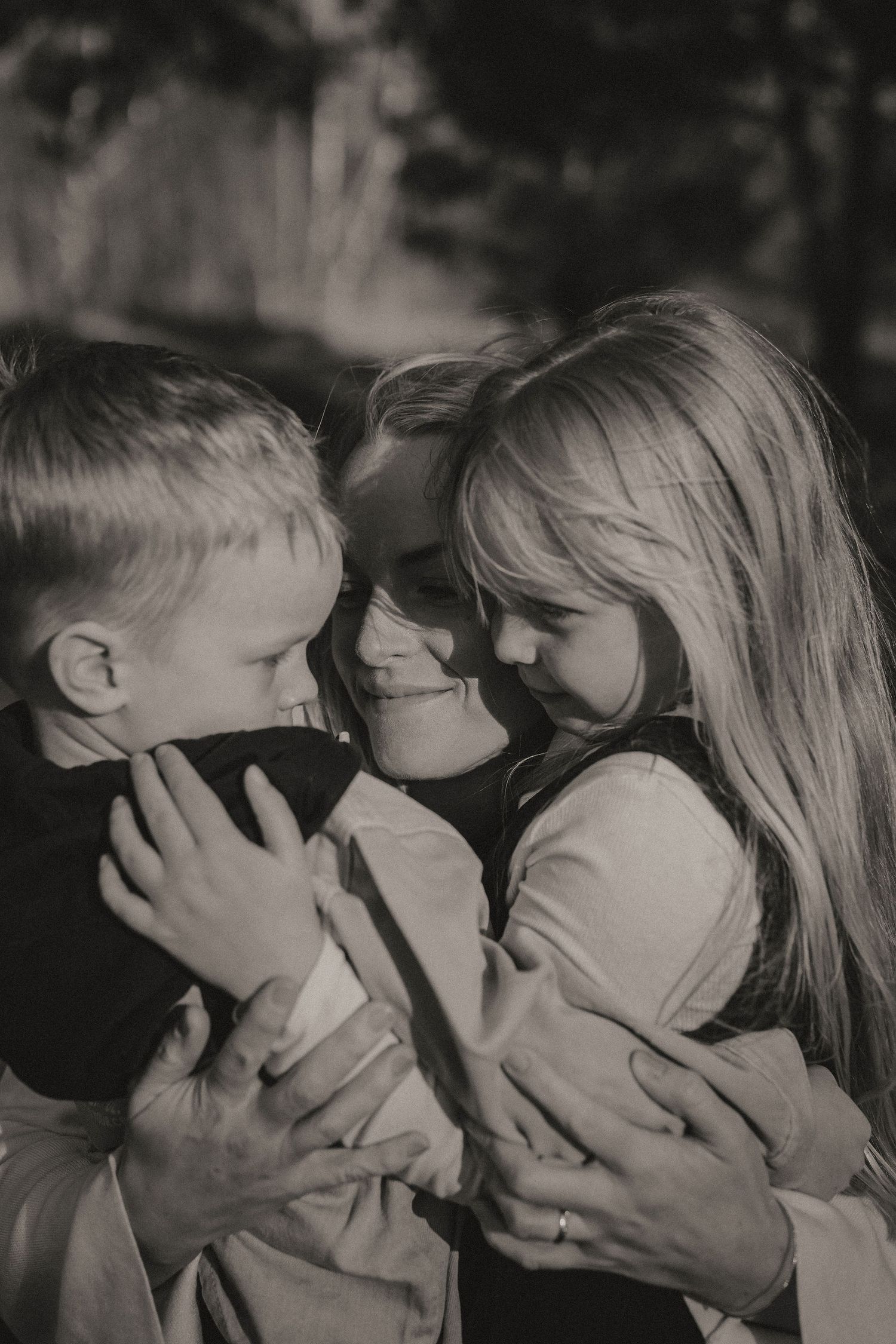 Black and white photograph showing intimate family moment with mother and children embracing and smiling together outdoors.