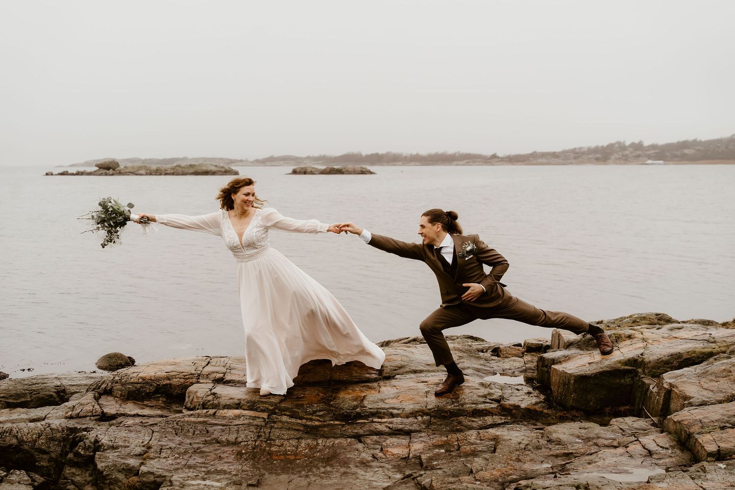 A couple in wedding attire playfully poses on rocky coastal shoreline at sunset with groom helping bride keep balance.