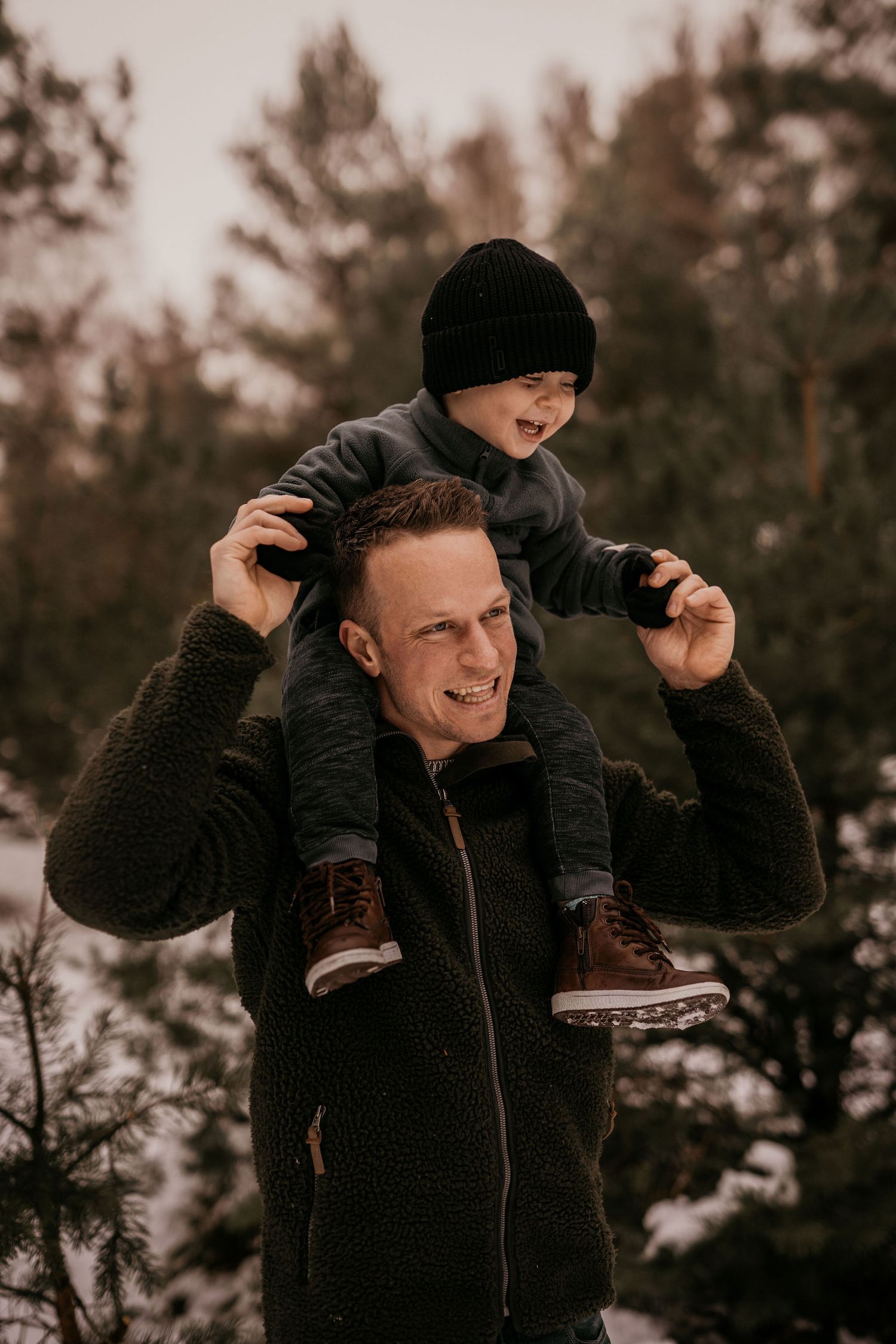 A child in a black winter hat sits on a parent's shoulders during a winter day in the woods.