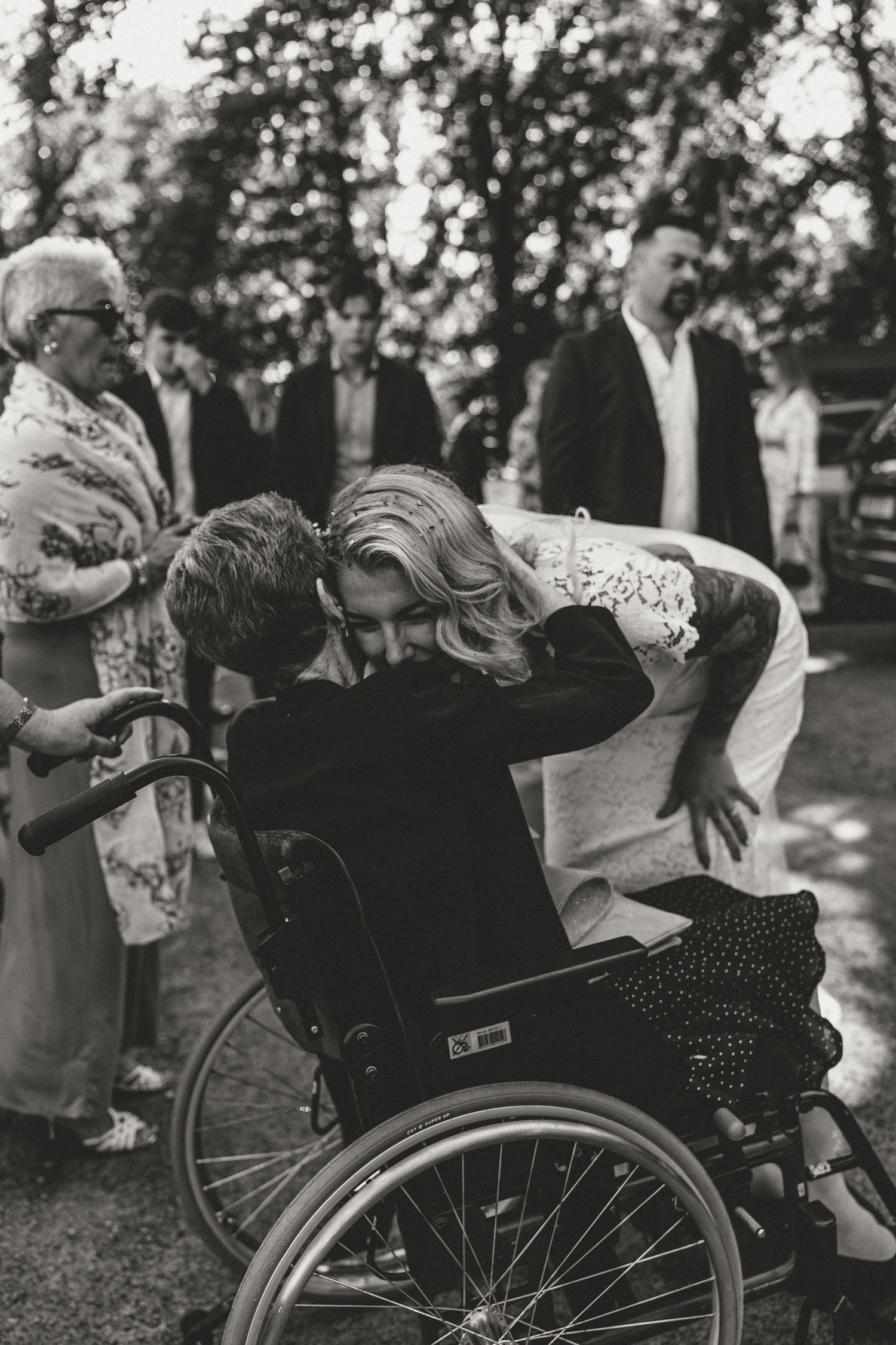 A tender moment captured in black and white as a bride leans over to embrace someone in a wheelchair during an outdoor ceremony.