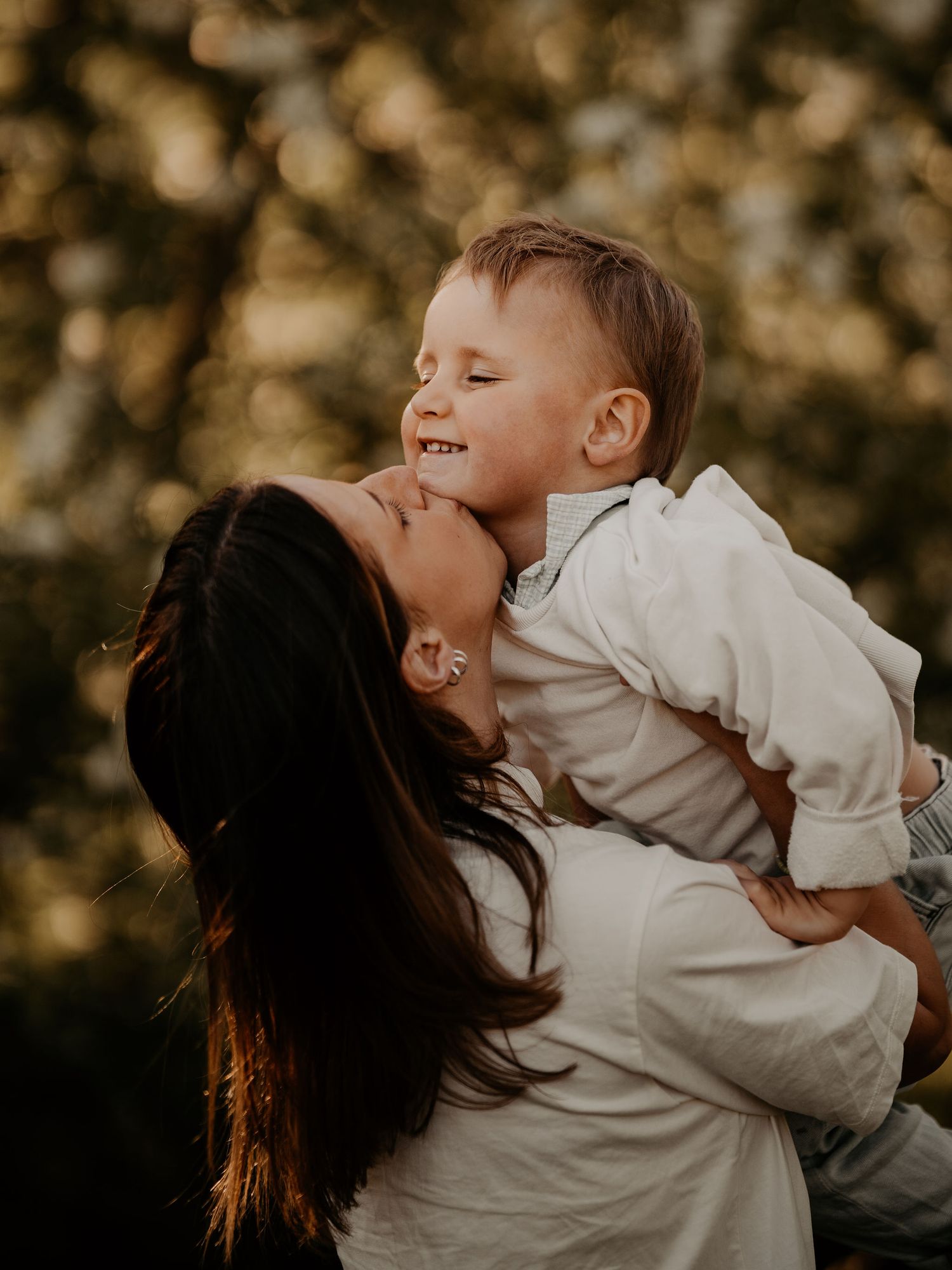 A tender moment captured at sunset as a woman in white embraces a joyful young child against a bokeh forest backdrop.