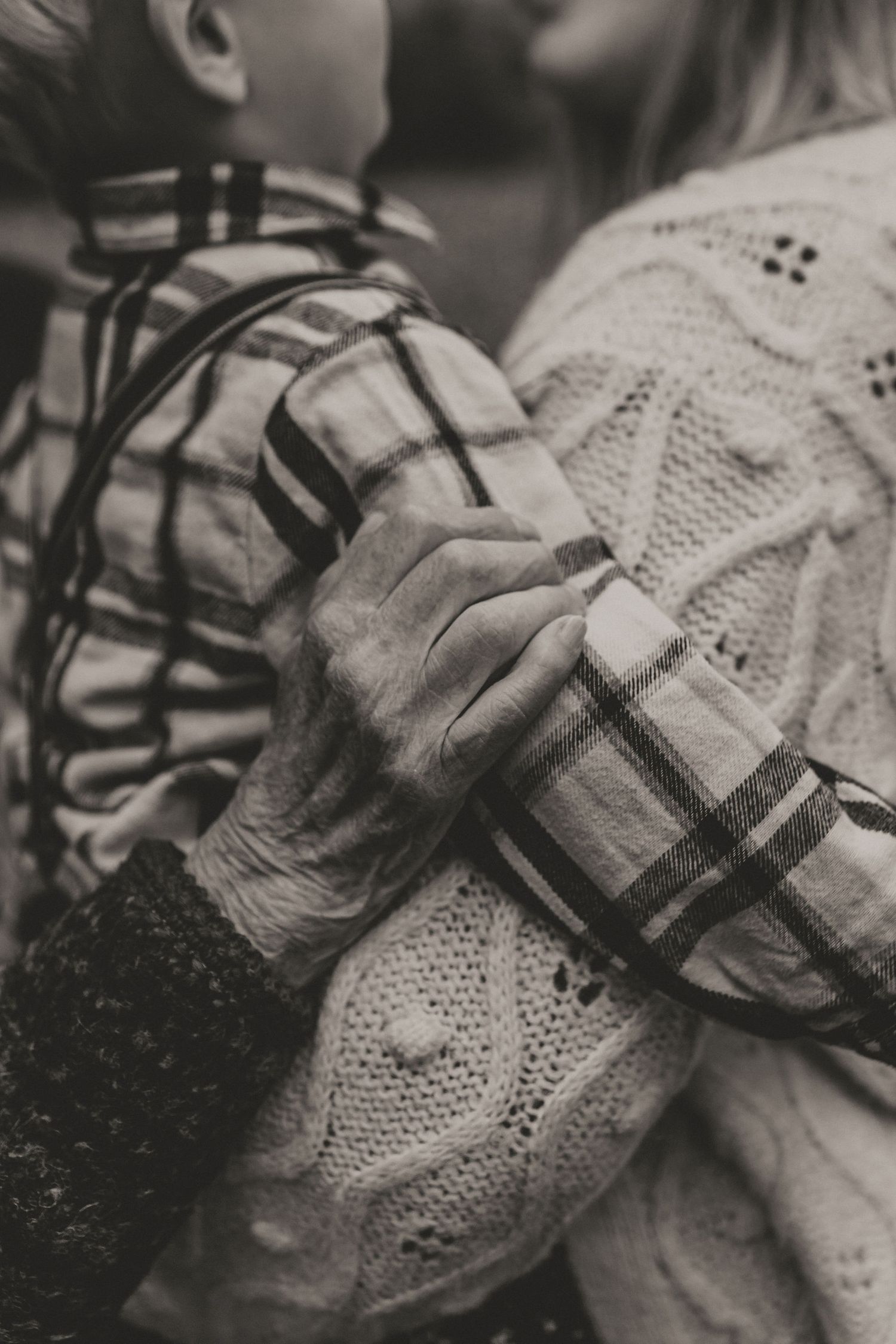 A close-up black and white photo of hands grasping a plaid shirt and knit sweater showing tender affection.