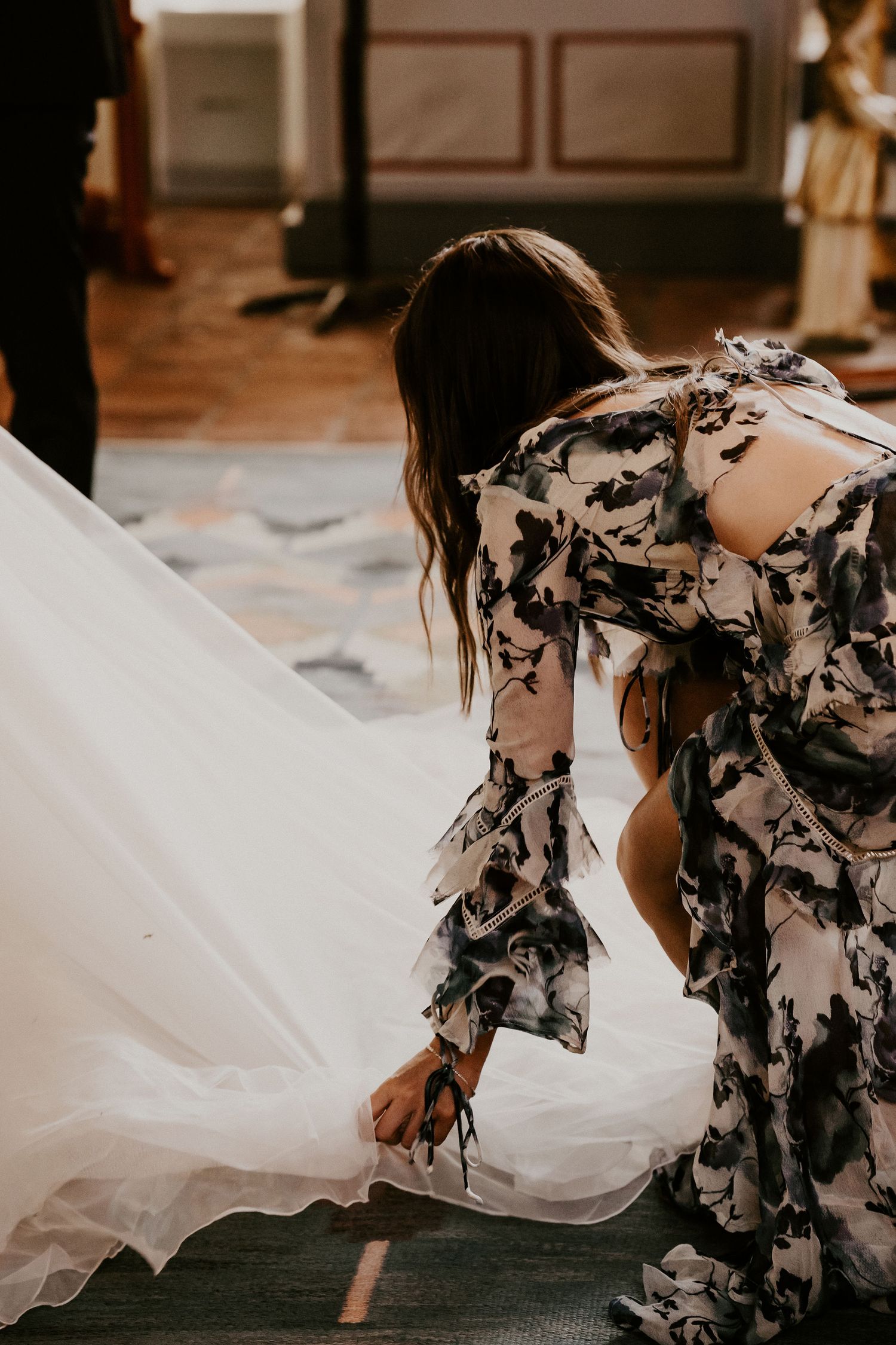 Someone wearing a floral print dress helps adjust white bridal gown hem during wedding preparations.