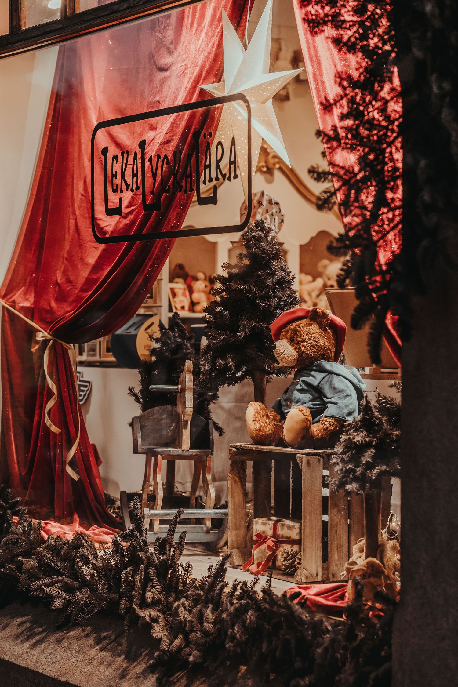 Cozy Christmas storefront display with red curtains, a lit star, teddy bears and a decorated tree in the window.