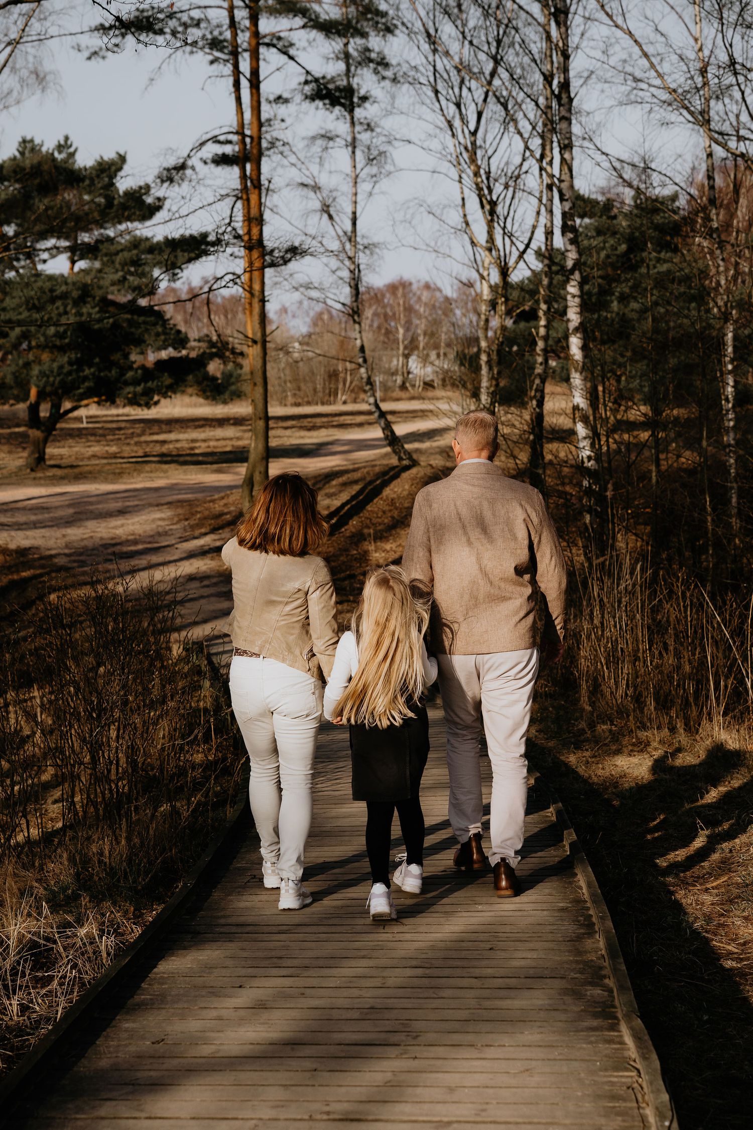 Three people walk together along a wooden boardwalk in nature during a sunny day wearing matching beige outfits.