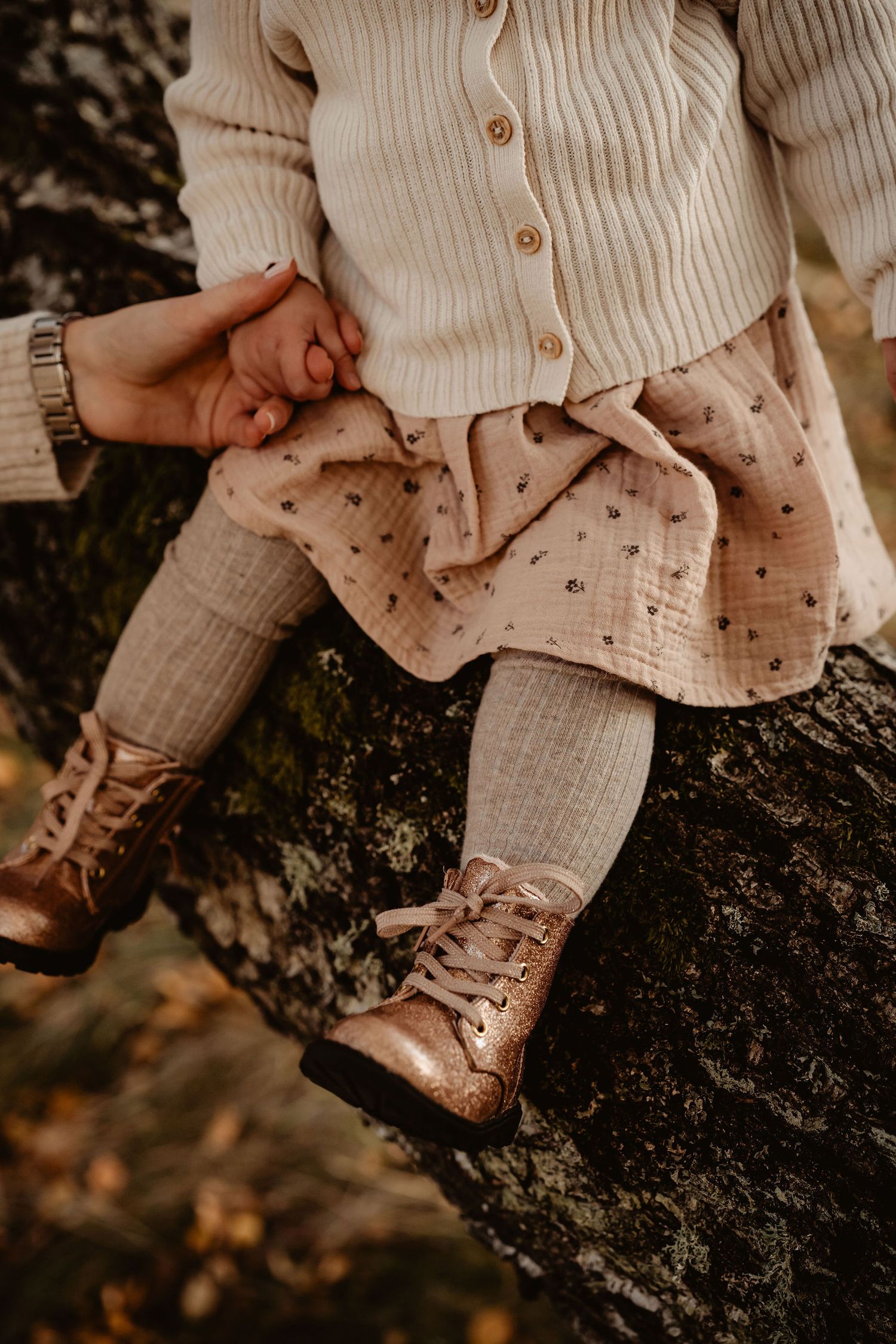 A child wearing cream ribbed cardigan, patterned skirt, gray tights and vintage brown leather lace-up boots sits on tree bark.