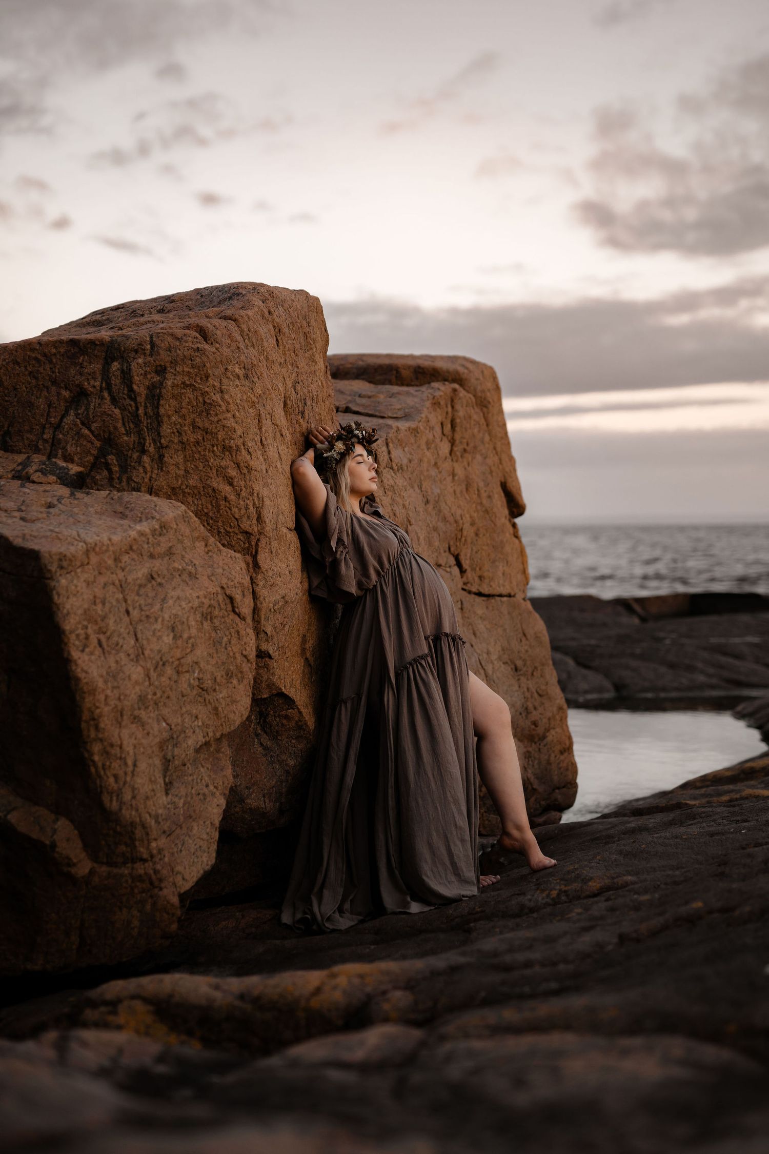 A figure in a flowing gray dress poses dramatically against rocky coastal cliffs at sunset.
