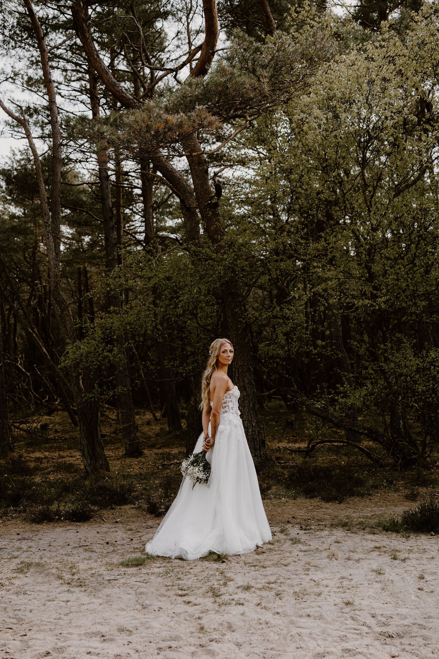 A wedding portrait of a figure in a flowing white gown standing on a sandy path surrounded by pine trees.