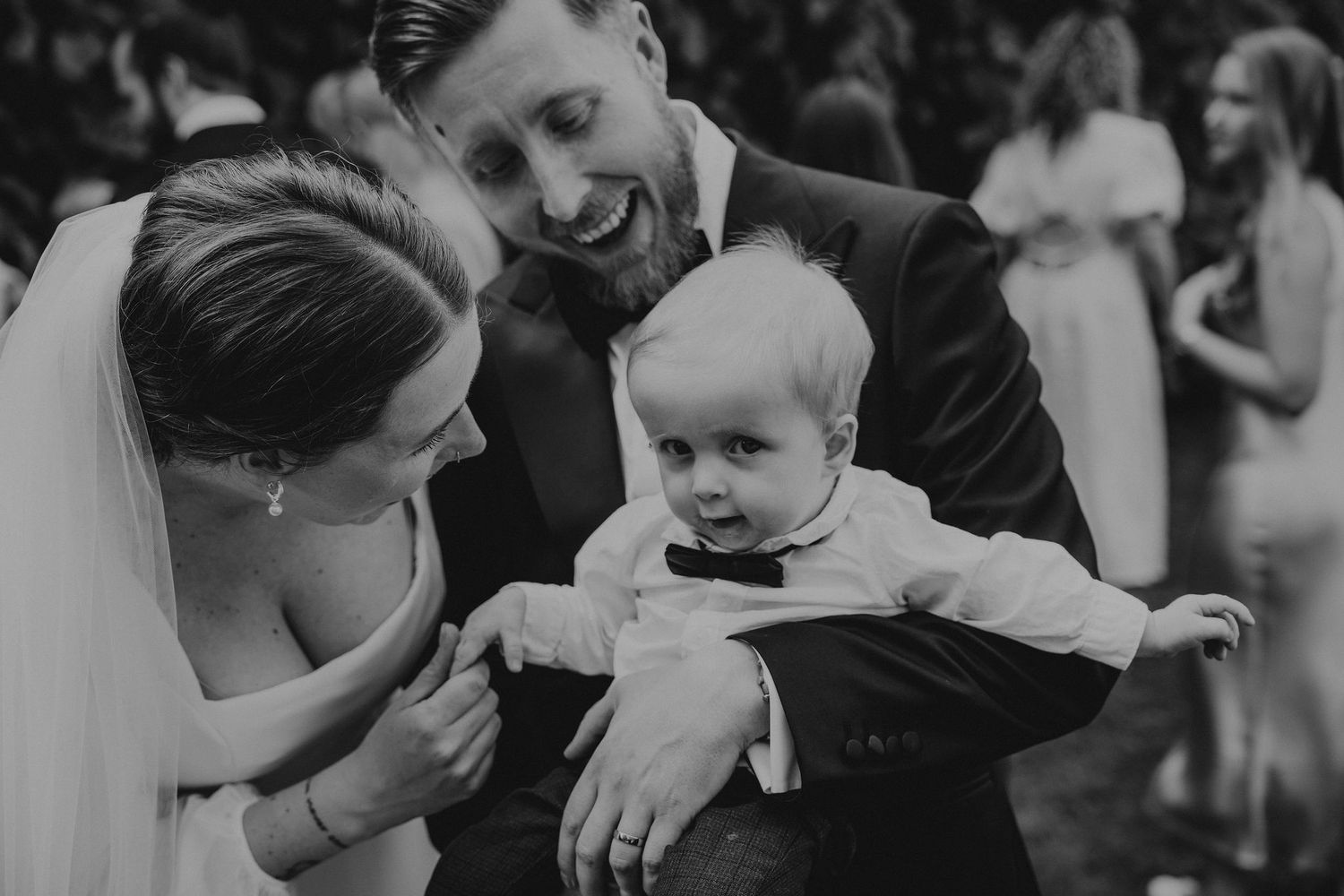 A tender black and white wedding moment shows a smiling couple interacting with a baby in formal attire.