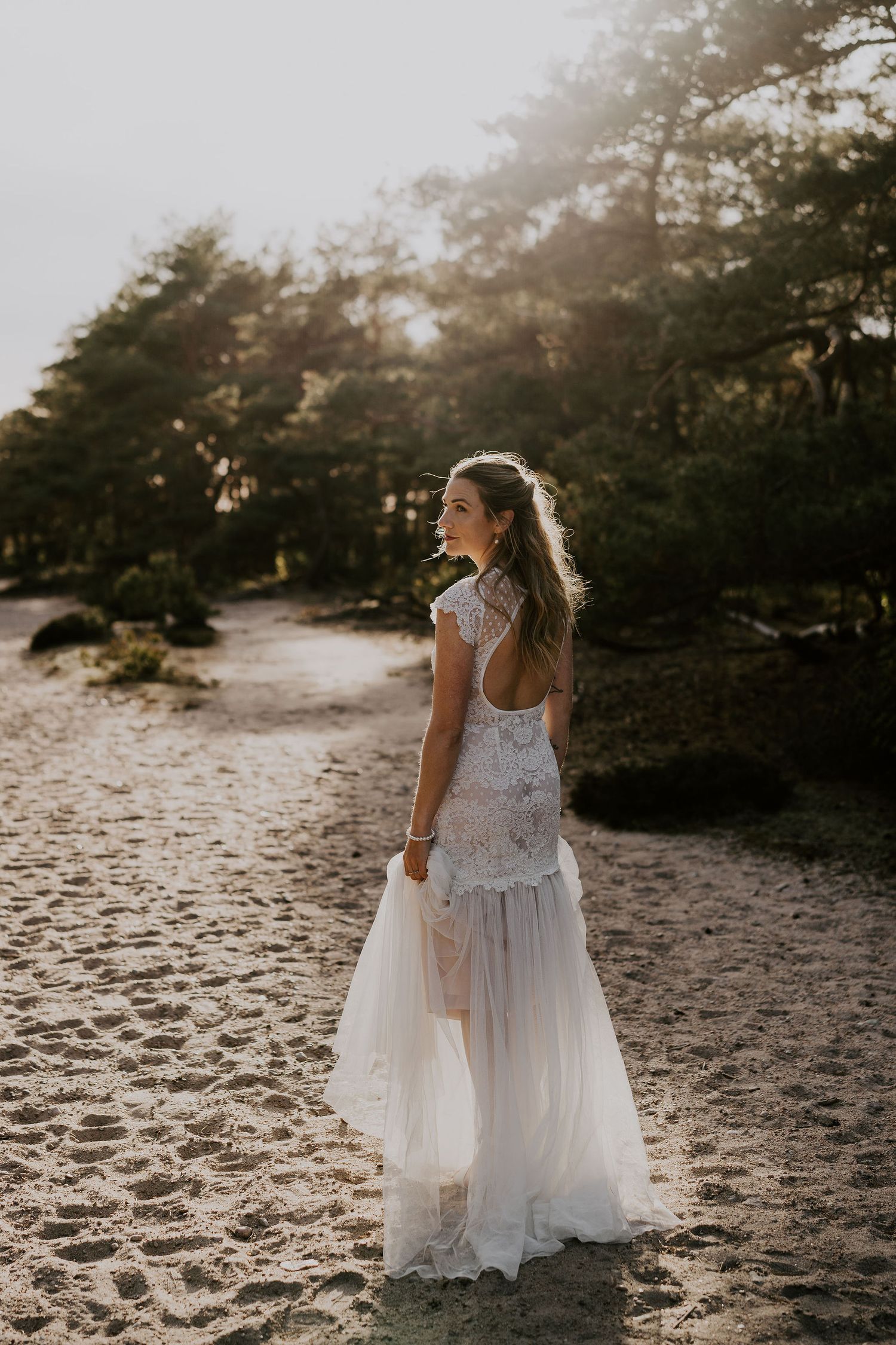 A bride in a flowing white lace and tulle wedding dress stands on a sandy beach path at golden hour.