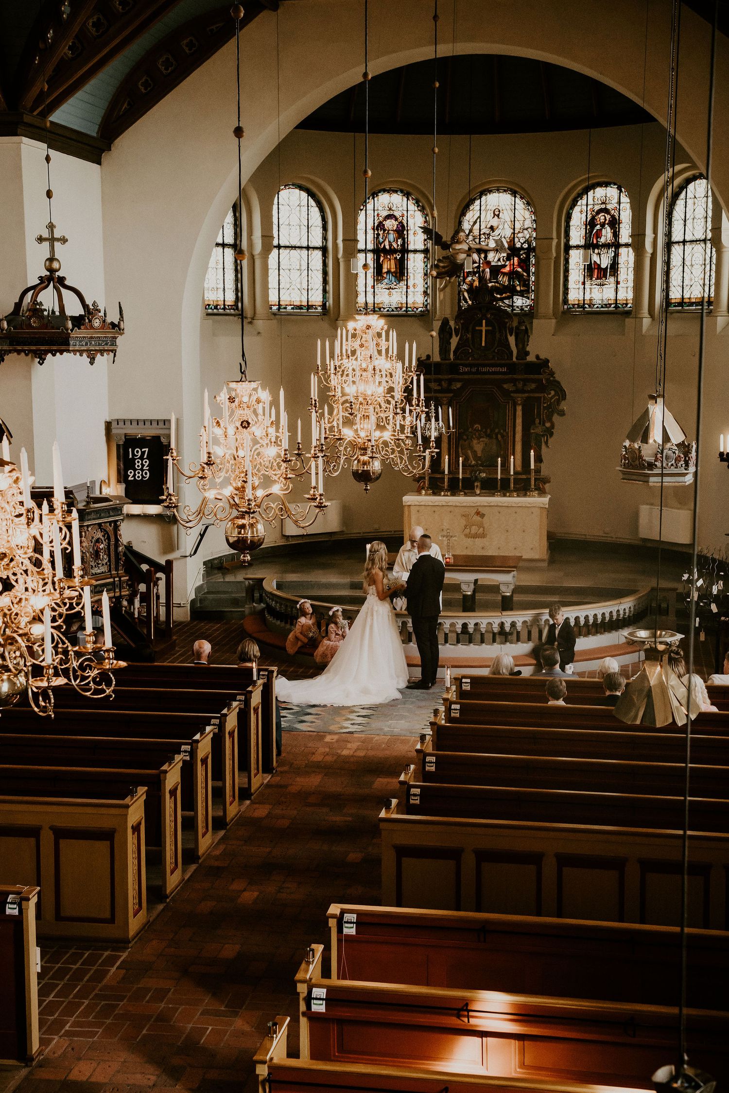 Elegant church wedding ceremony with ornate crystal chandeliers and arched stained glass windows in warm vintage tones.