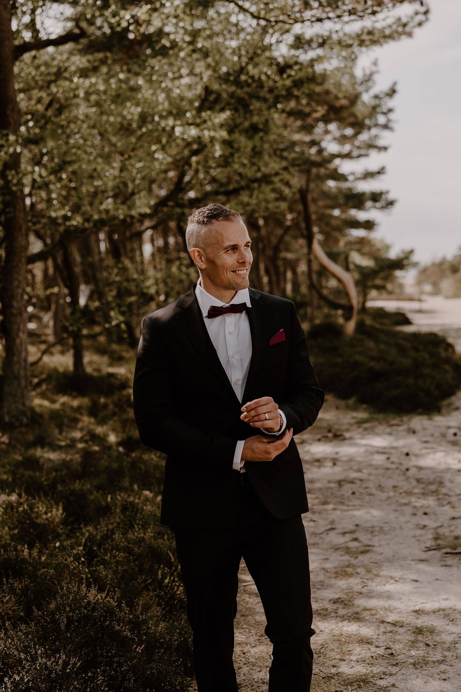 A figure in a classic black tuxedo with bow tie and red boutonniere stands on a forest path in warm sunlight.