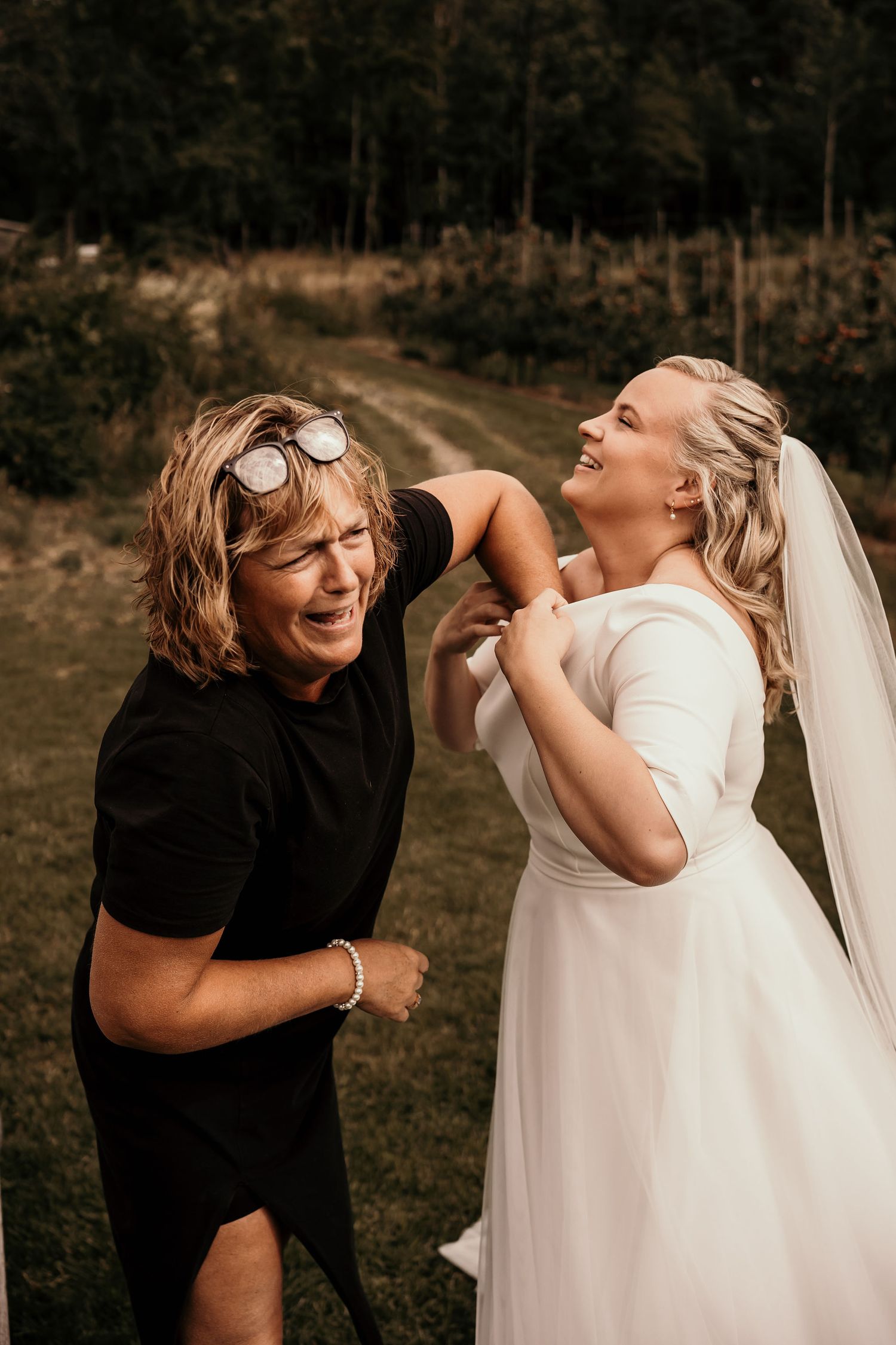 Two people share a candid joyful moment outdoors, one wearing black and one in a white wedding dress with veil.